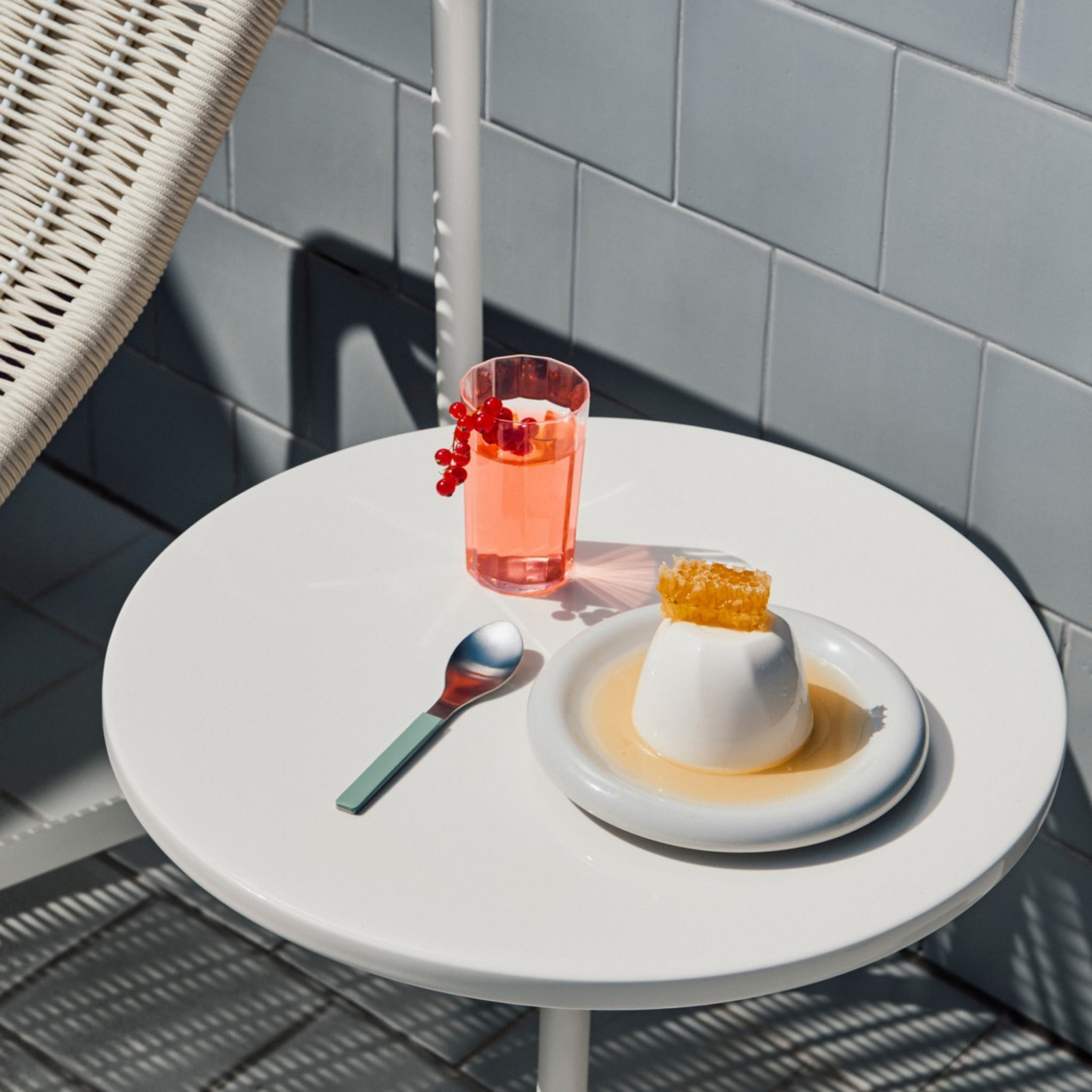 Op een witte ronde tafel liggen panna cotta met honingraat op een bord, een lepel en een roze drank met rode bessen in Hay Angle Longdrink Glazen van Hay. Zonlicht werpt schaduwen over het tafereel en de betegelde muur op de achtergrond.