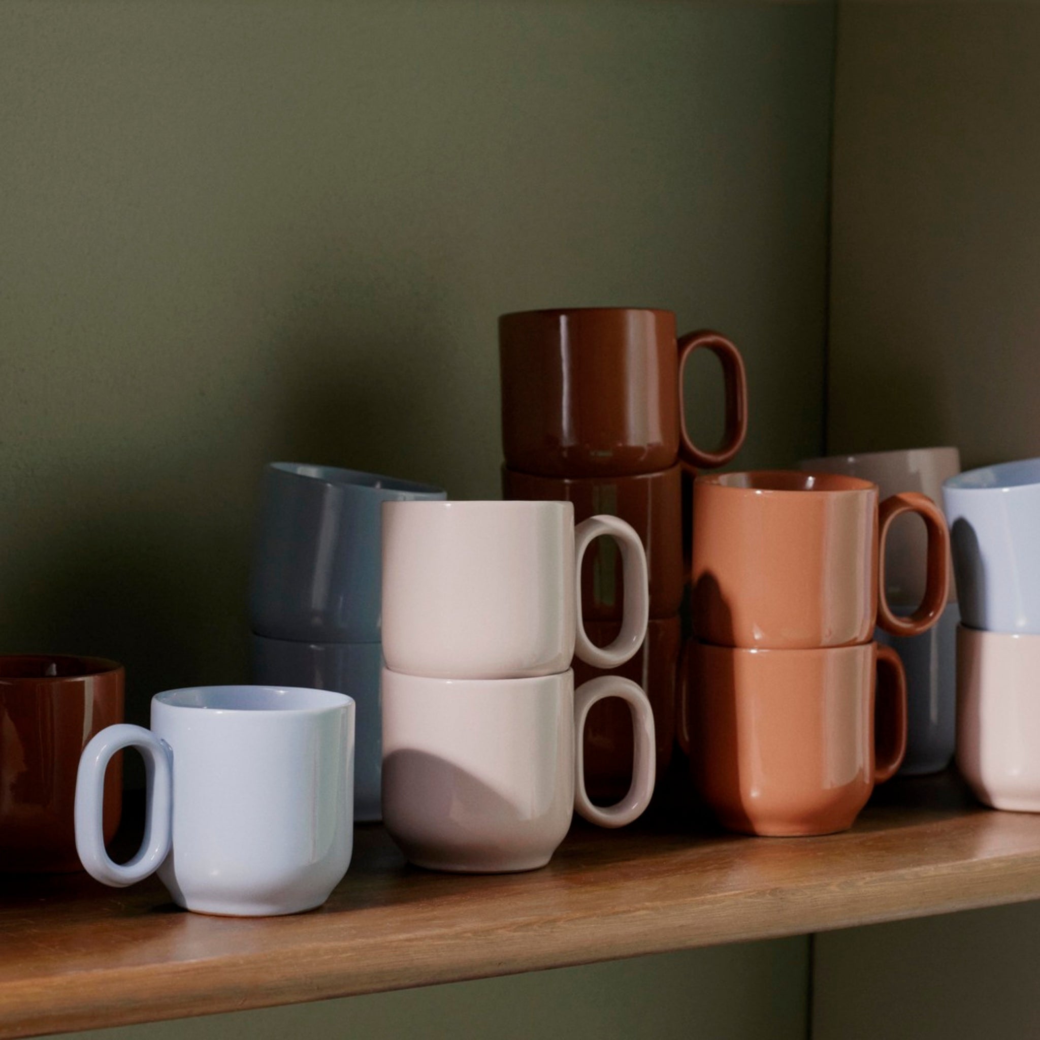 Een set Hay Barro Bekers Roze 2 st. keramische mokken in zachte pastel- en aardetinten, zoals lichtblauw, beige en bruin, netjes gestapeld op een houten plank tegen een gedempte groene muur.