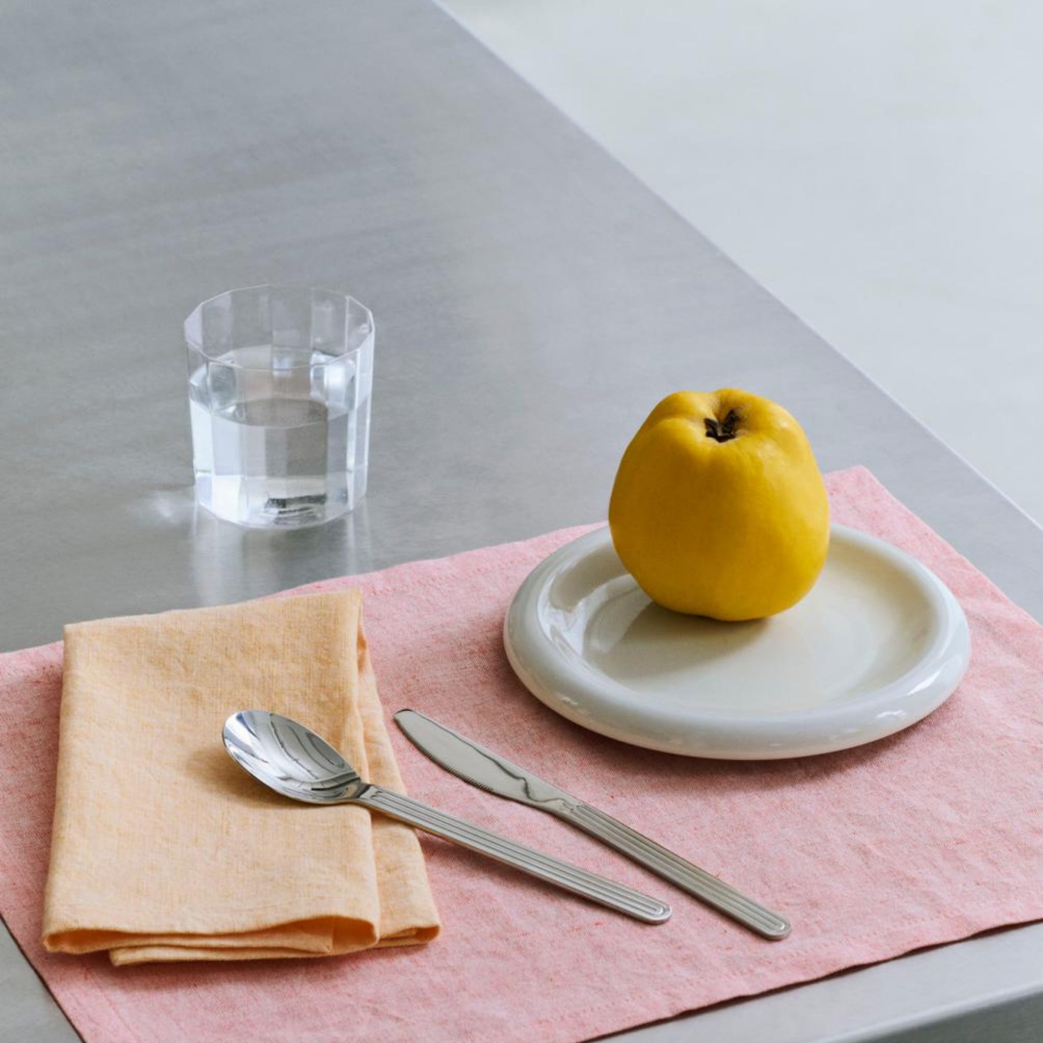 Een gele appel op een wit bord rust op een roze placemat, naast een perzikkleurig servet met bestek en een kristallijn glas water van Hay Angle Waterglazen 4 st. dat elegantie toevoegt aan de metallic tafel.