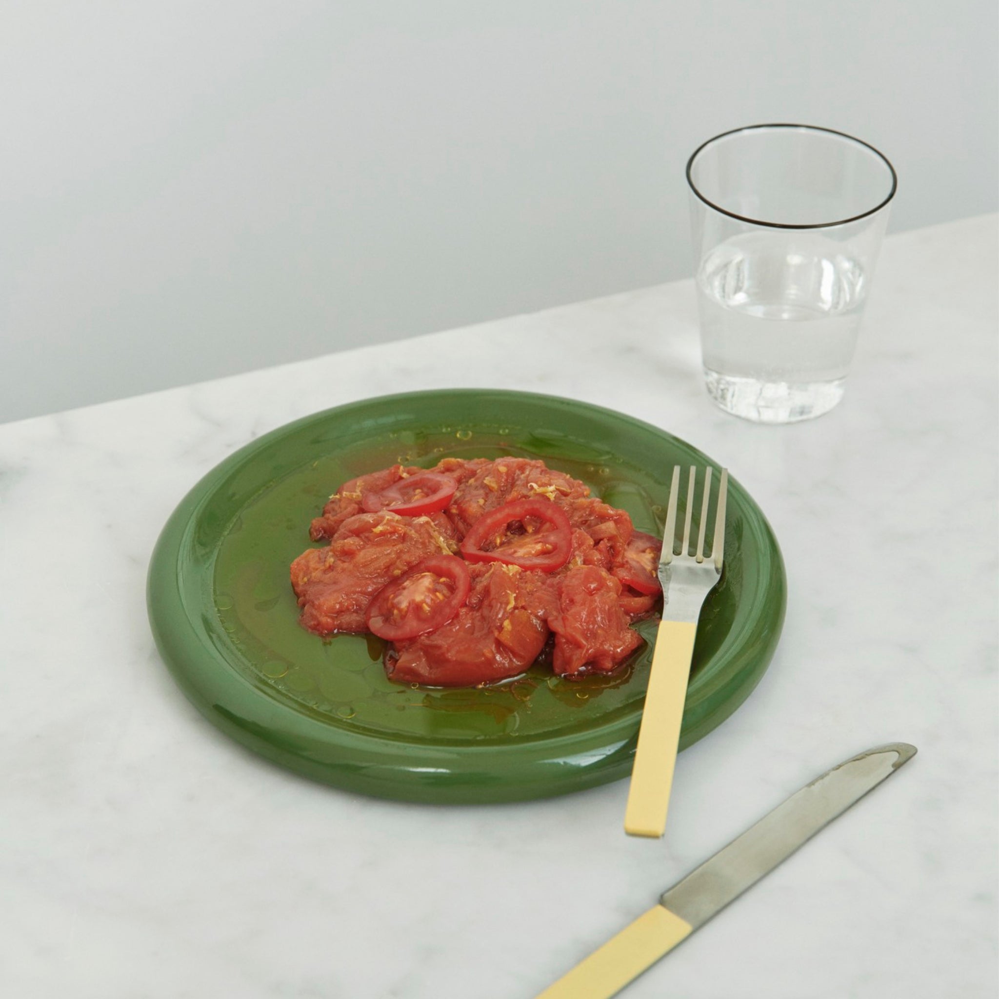 Gesneden tomaten met olie worden geserveerd op een Hay Barro Borden Groen D24 bord van Hay, met een vork met gele handgreep, plus een mes en een glas water in de buurt op een wit marmeren oppervlak.
