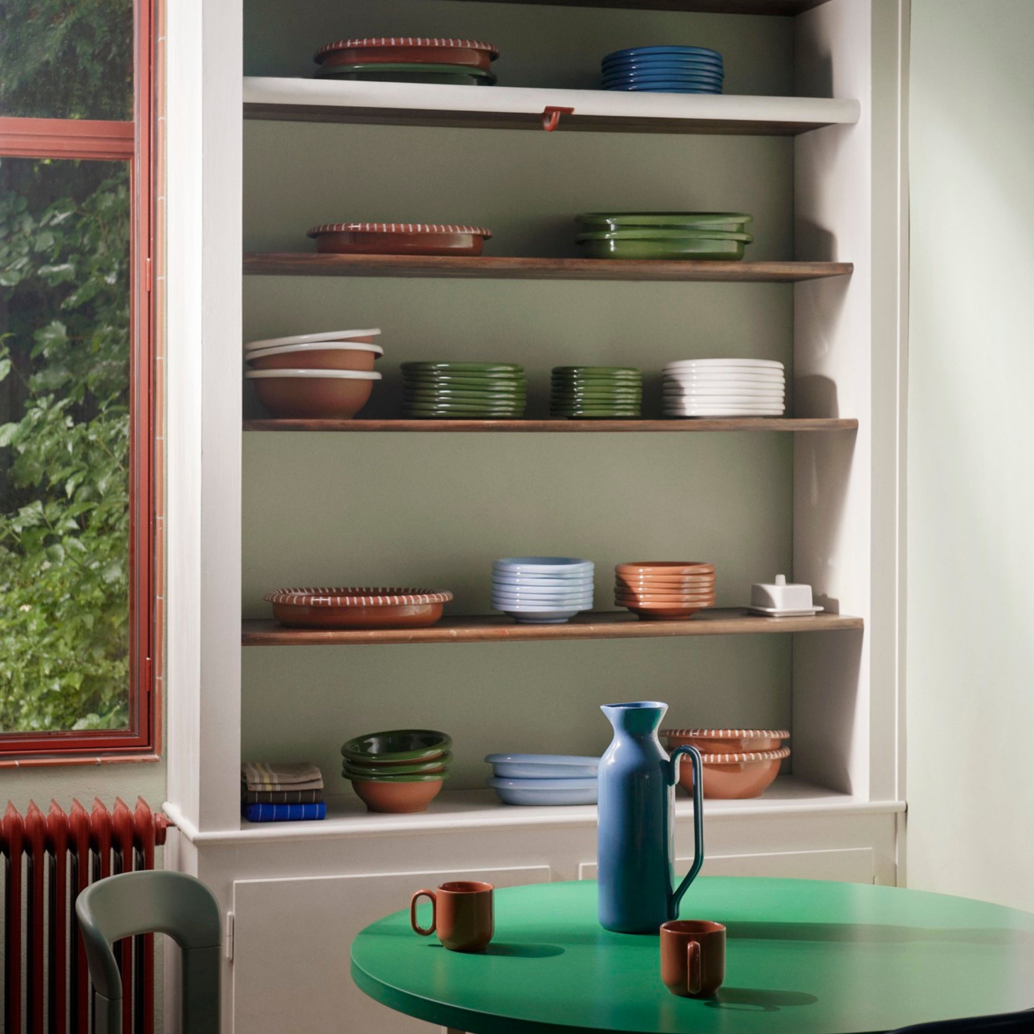 Op open planken staan netjes gestapelde Hay Barro Borden Groen D18 2 st. van Hay, samen met terracotta servies in groen, bruin, wit en blauw. Een ronde groene tafel met twee mokken en een blauwe kan staat bij een rood omlijst raam met uitzicht op groen.
