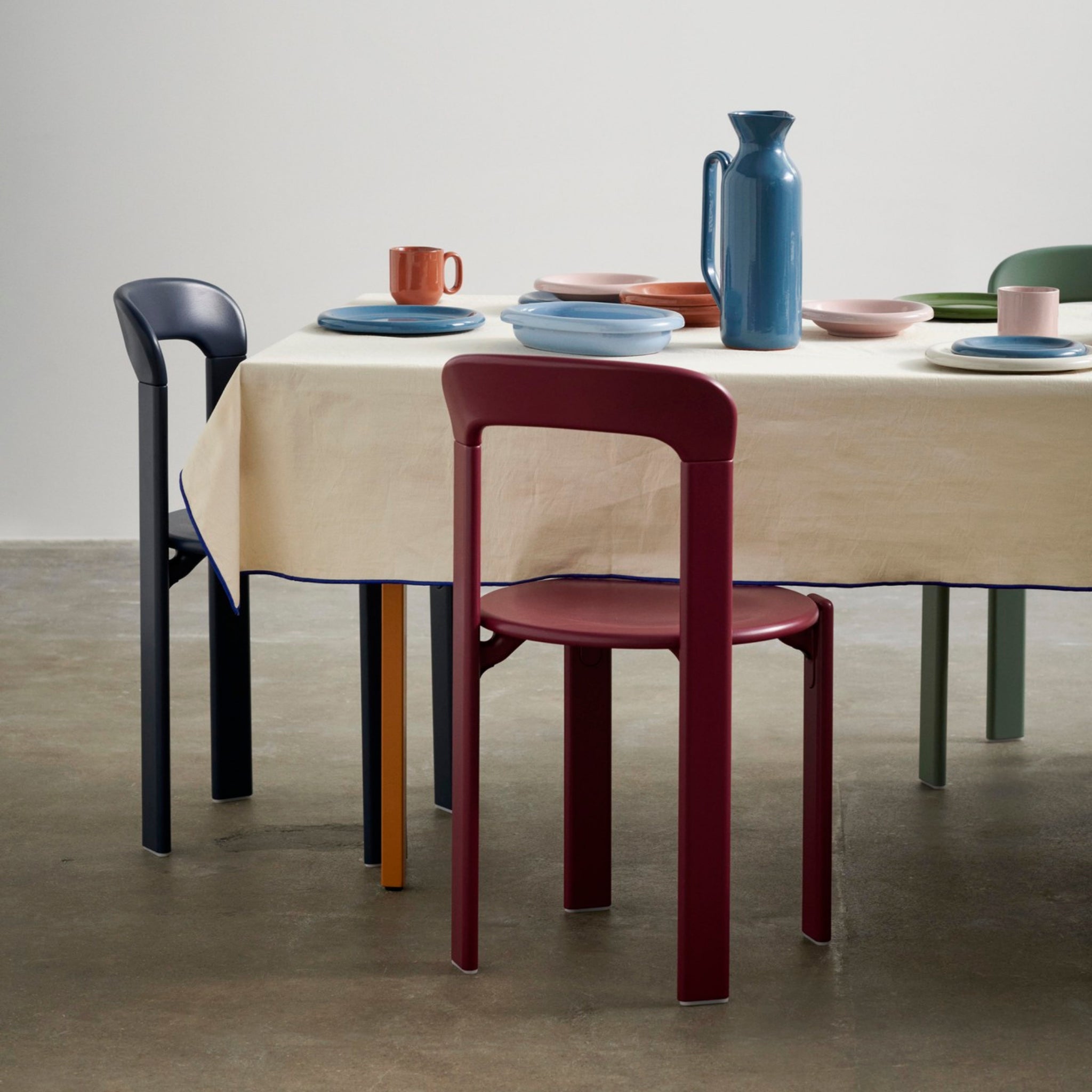 Een ronde tafel met een crèmekleurig tafelkleed is gedekt met kleurrijke borden, Hay Barro Bekers Roze 2 st. van Hay, en een blauwe kan, omringd door mismatched moderne stoelen in rood, marineblauw, groen en geel op een betonnen vloer.