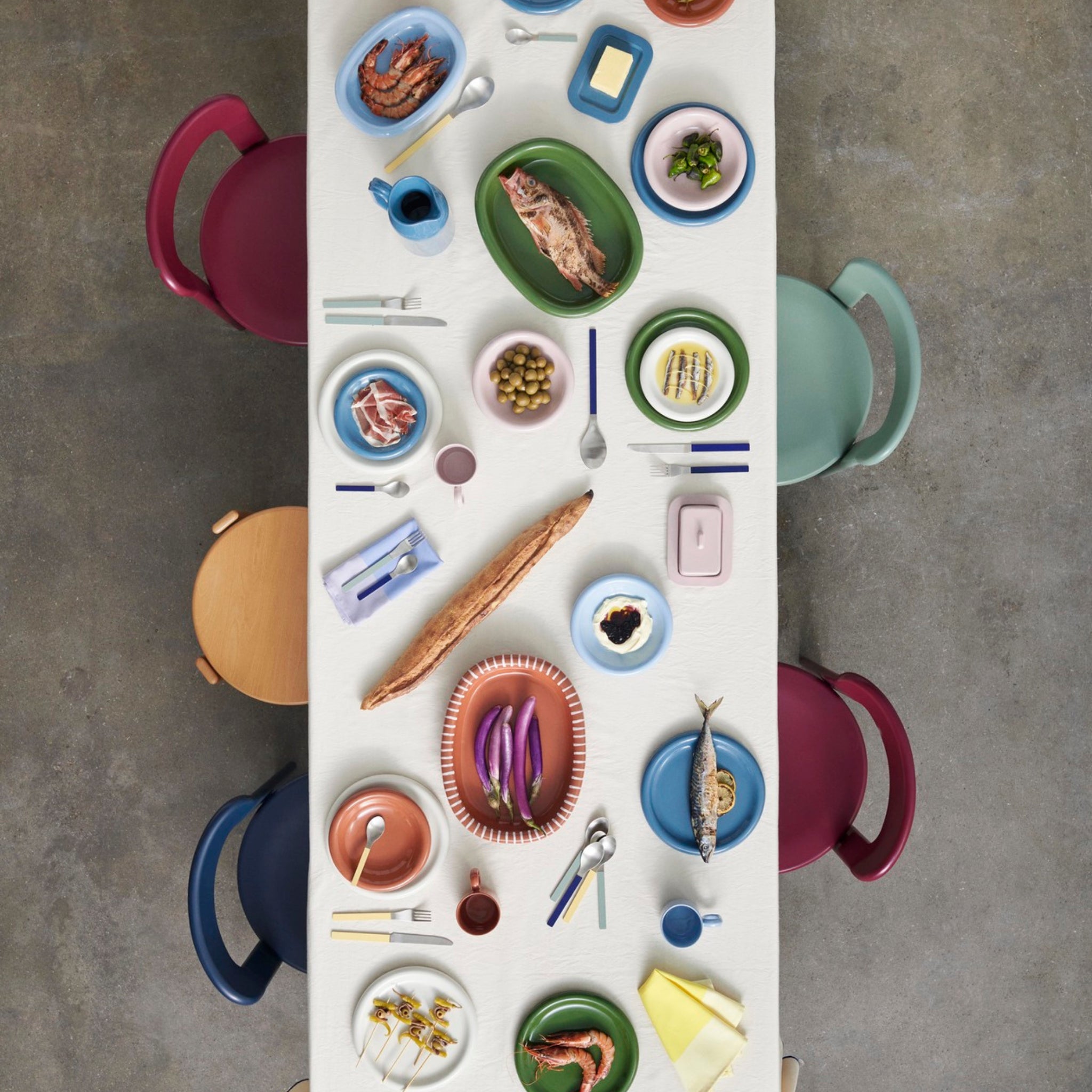 Een bovenaanzicht van een eettafel gedekt met Hay Barro Borden Groen D18 borden van Hay, geaccentueerd met terracotta borden, kleurrijk eten zoals vis, garnalen en groenten, en vier heldere stoelen op een grijze vloer.