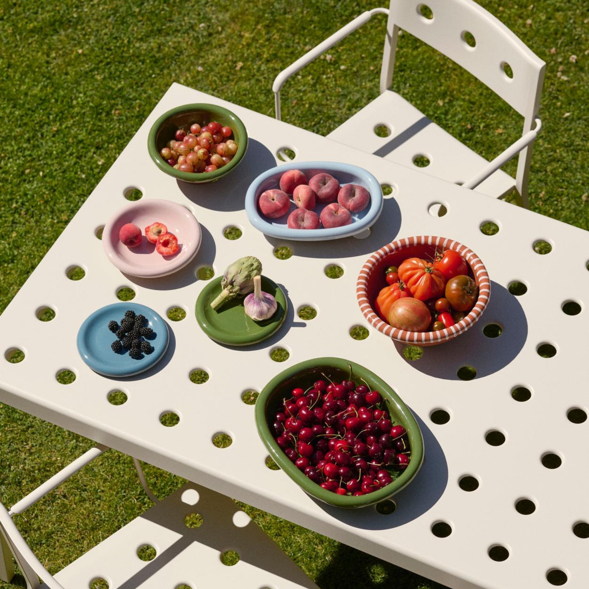 Op het gras staat een witte geperforeerde tafel met bijpassende stoelen met daarop de Hay Barro Ovale Schaal Lichtblauw 20/27 gevuld met druiven, perziken, kersen, bramen, een artisjok en heirloom tomaten.