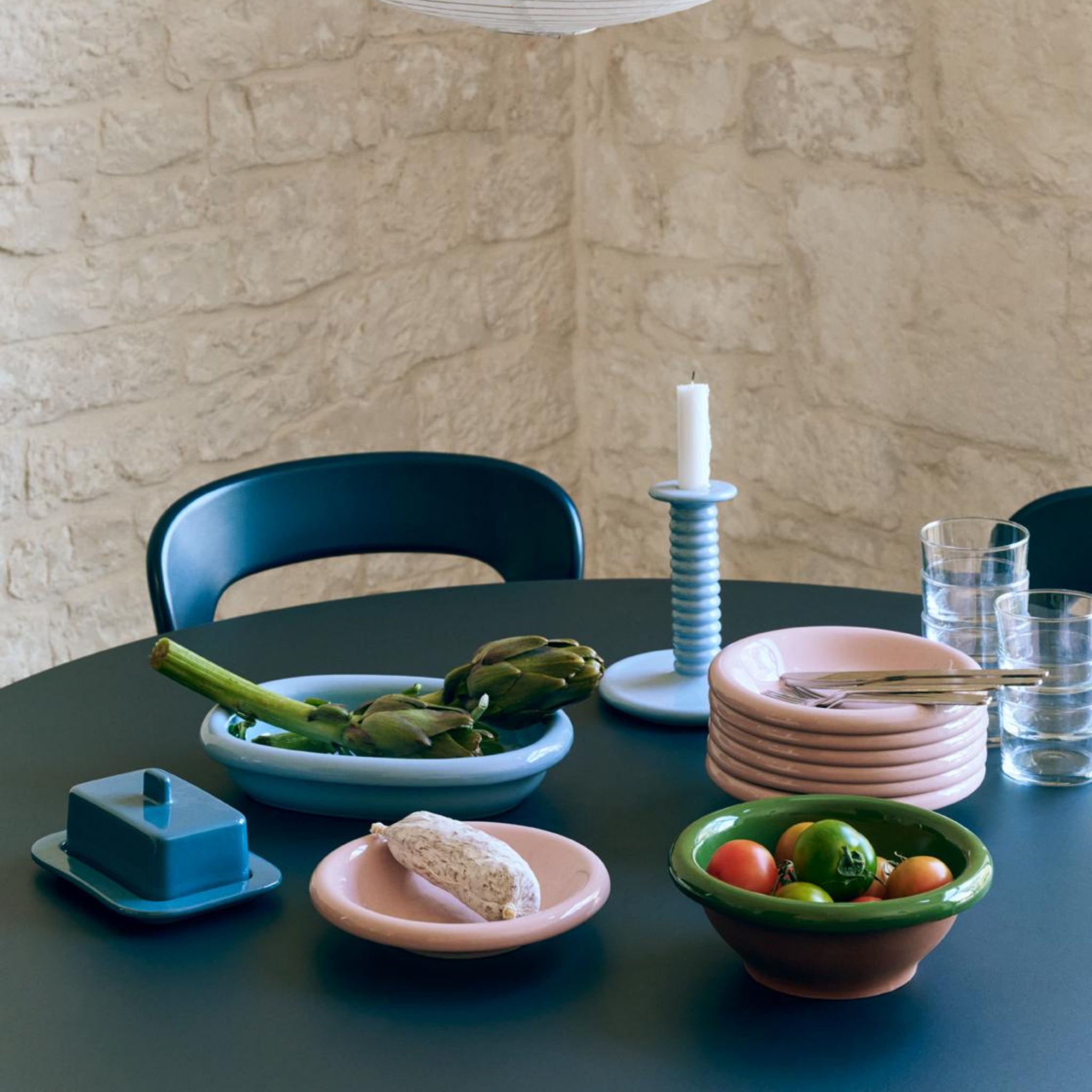 Een ronde tafel gedekt met pastelkleurige keramieken schaaltjes, een botervloot, Portugees servies, groenten in de Hay Barro Kom Groen D21 van Hay, glazen kopjes en een hoge kaars staat tegen een lichte stenen muur en donkere stoel.