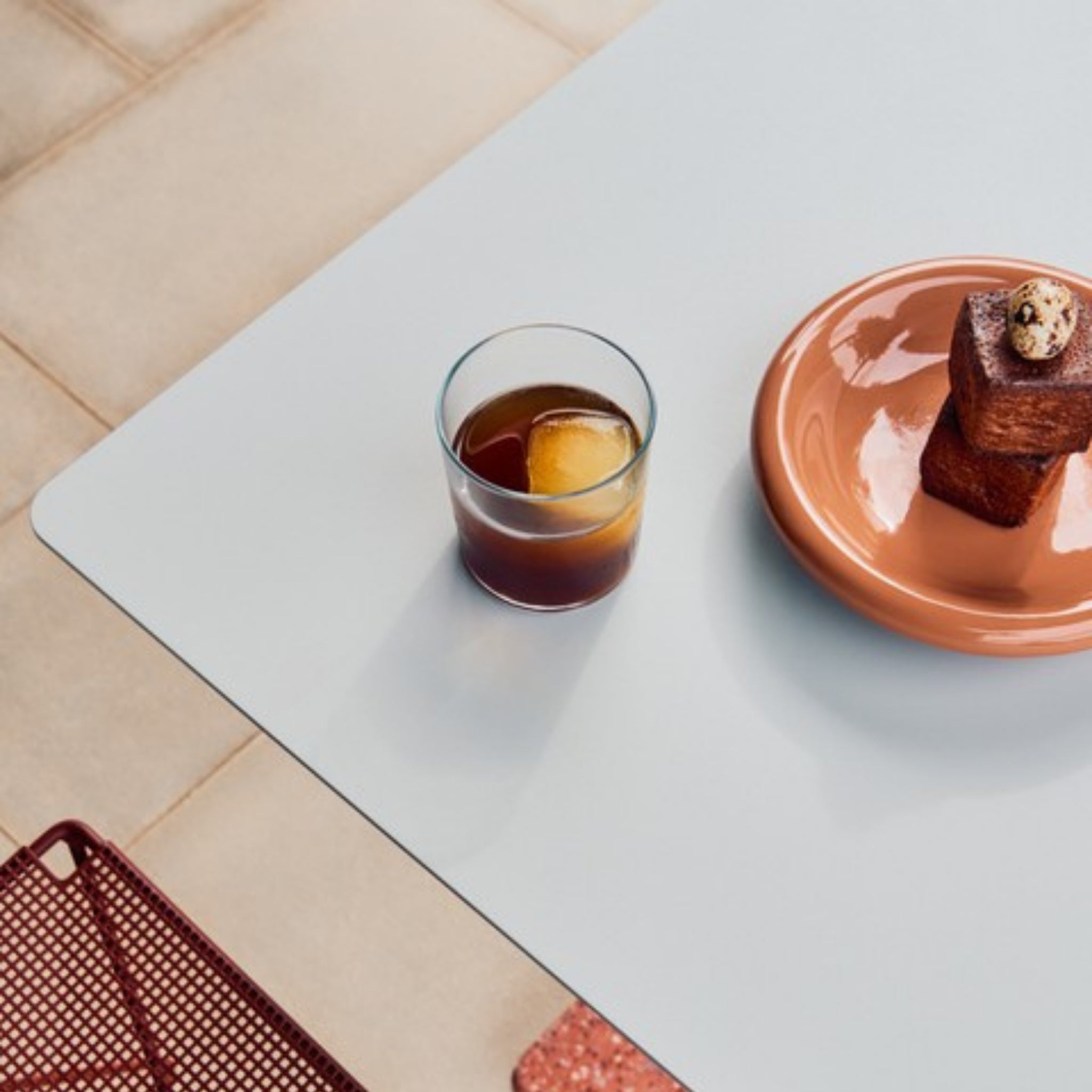 Een glas donkere ijskoffie met een groot ijsblokje staat naast een Hay Barro Diepe Borden Terracotta D20 2 st. bord van Hay, met daarop gebakjes met een kwarteleitje, op een lichtblauwe tafel.