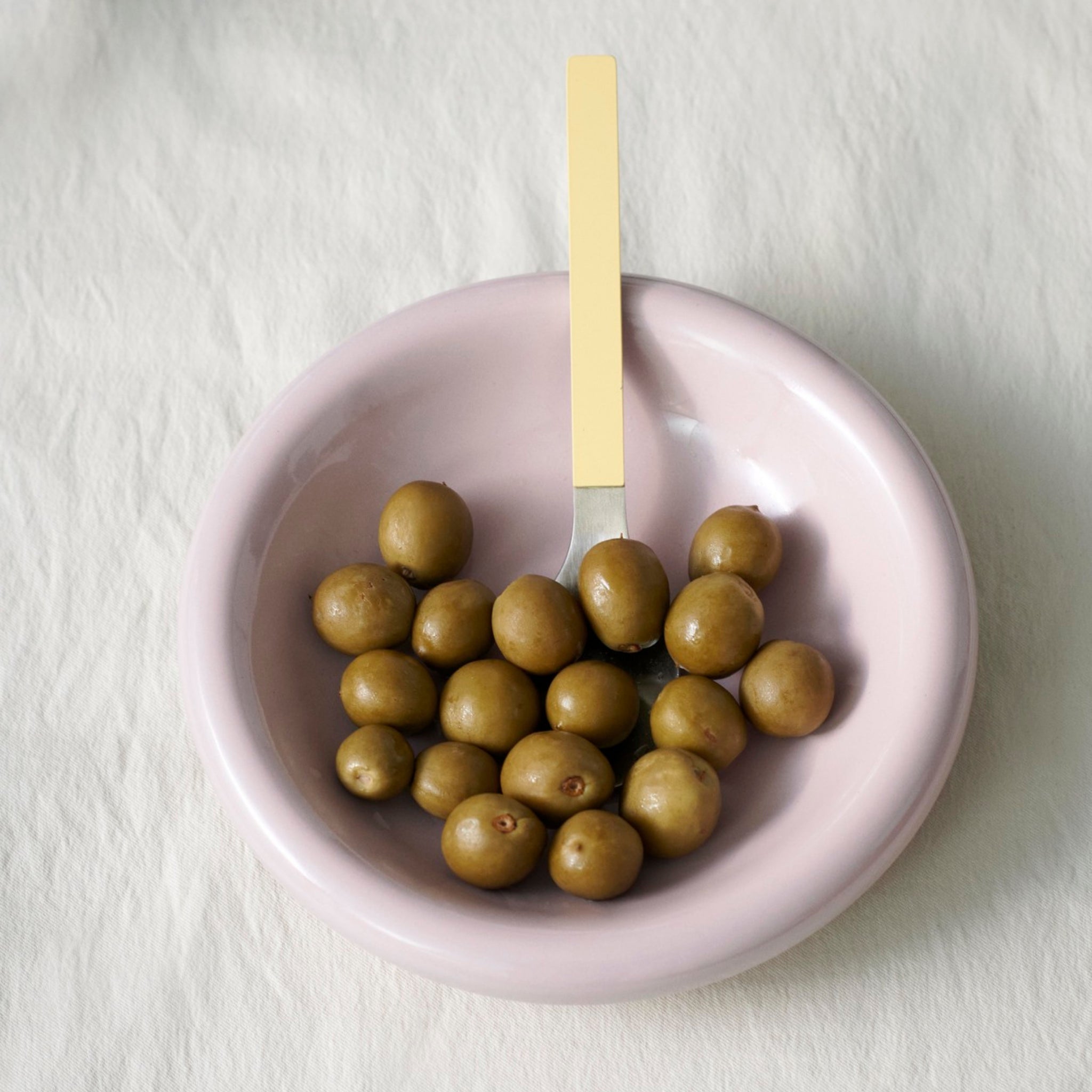 Een Hay Barro Diepe Borden Roze D20 schaal uit Hay, gevuld met groene olijven, staat op een wit kleed. Een vork met een lichtgeel handvat rust in de roze kom tussen de olijven.