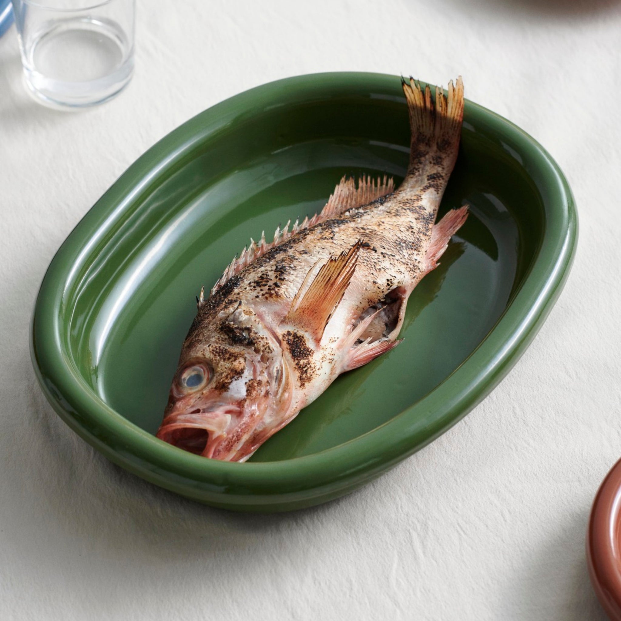 Een hele rauwe vis met open bek ligt in een Hay Barro Ovale Schaal Groen 27/36 van Hay, onderdeel van de terracotta collectie, op een wit tafelkleed met wat water; vlakbij staan een glas en een bord, die de elegante stijl van Rui Pereira oproepen.