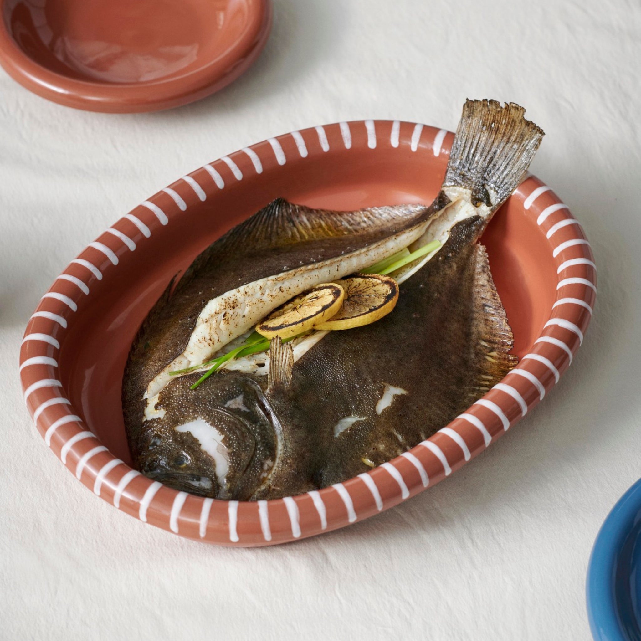 Een hele gekookte platvis gegarneerd met gegrilde citroen en groene uien rust in de Hay Barro Ovale Schaal Terracotta 27/36 van Hay, zijn roodbruine ovale vorm met witte randstrepen elegant gedekt op een wit tafelkleed.