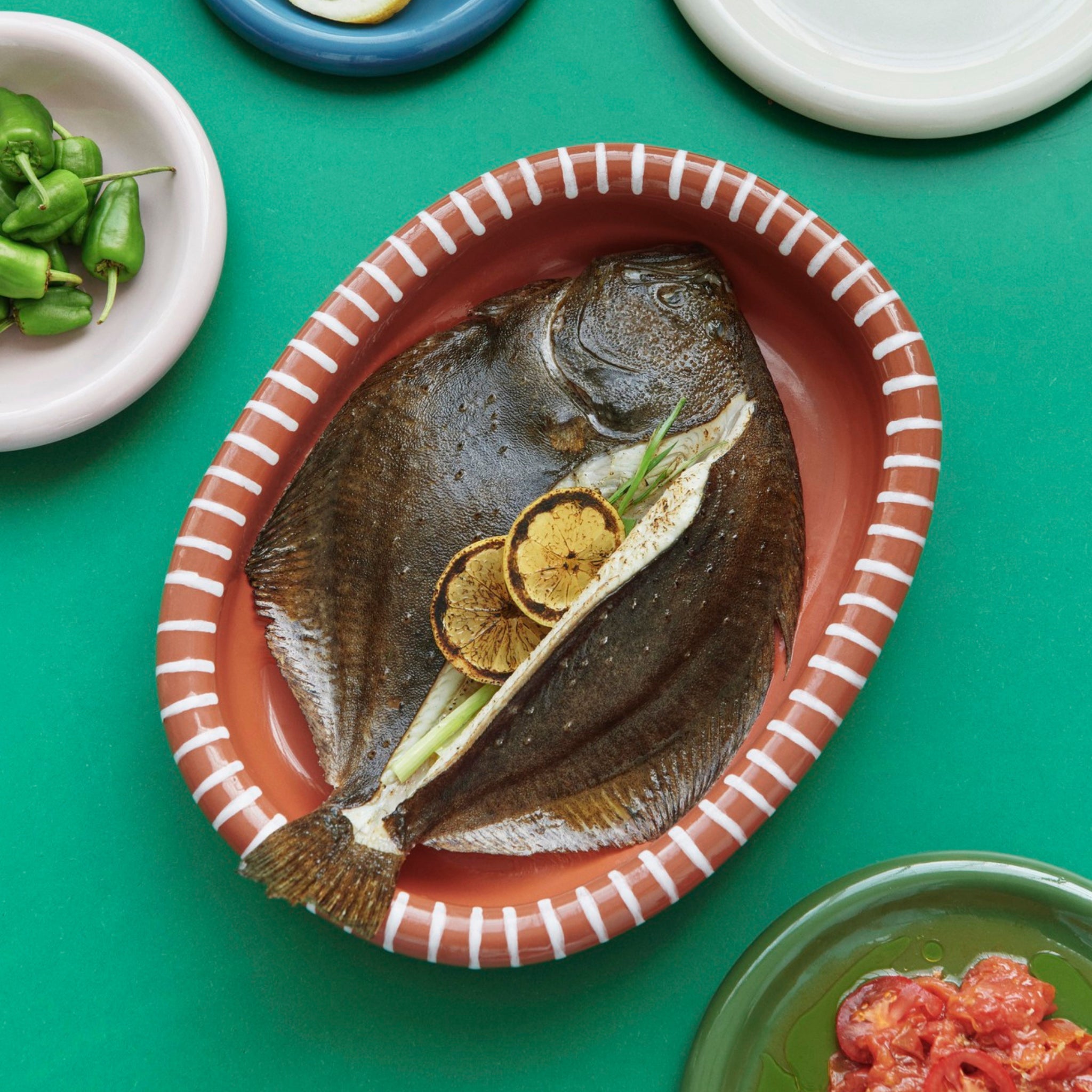 Een hele gekookte platte vis met gegrilde citroen en groene ui wordt geserveerd in een Hay Barro Ovale Schaal Terracotta 27/36 van Hay, omringd door kleine handbeschilderde schaaltjes op een groene tafel.