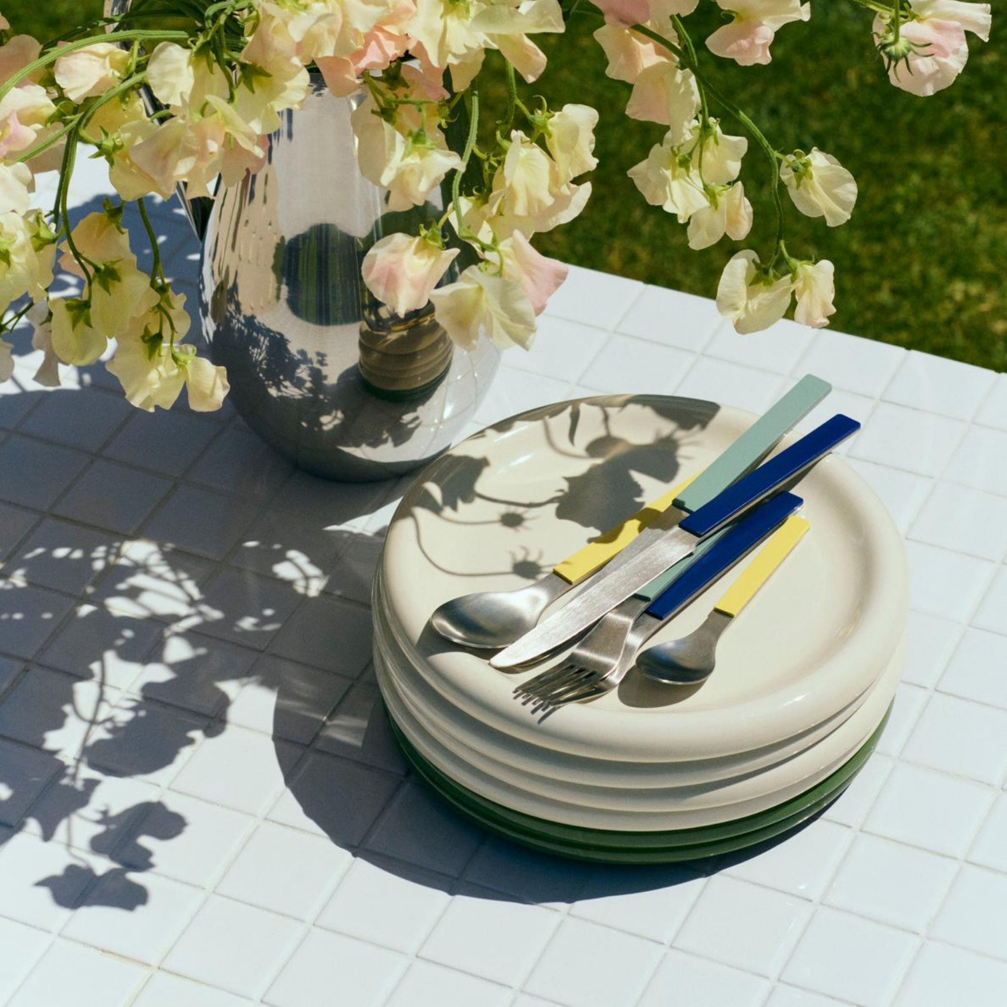 Een stapel Hay Barro Borden Off-White D24 borden van Hay staat op een zonovergoten betegelde buitentafel naast kleurrijk bestek en een vaas met lichtgele bloemen, die duidelijke schaduwen werpen.