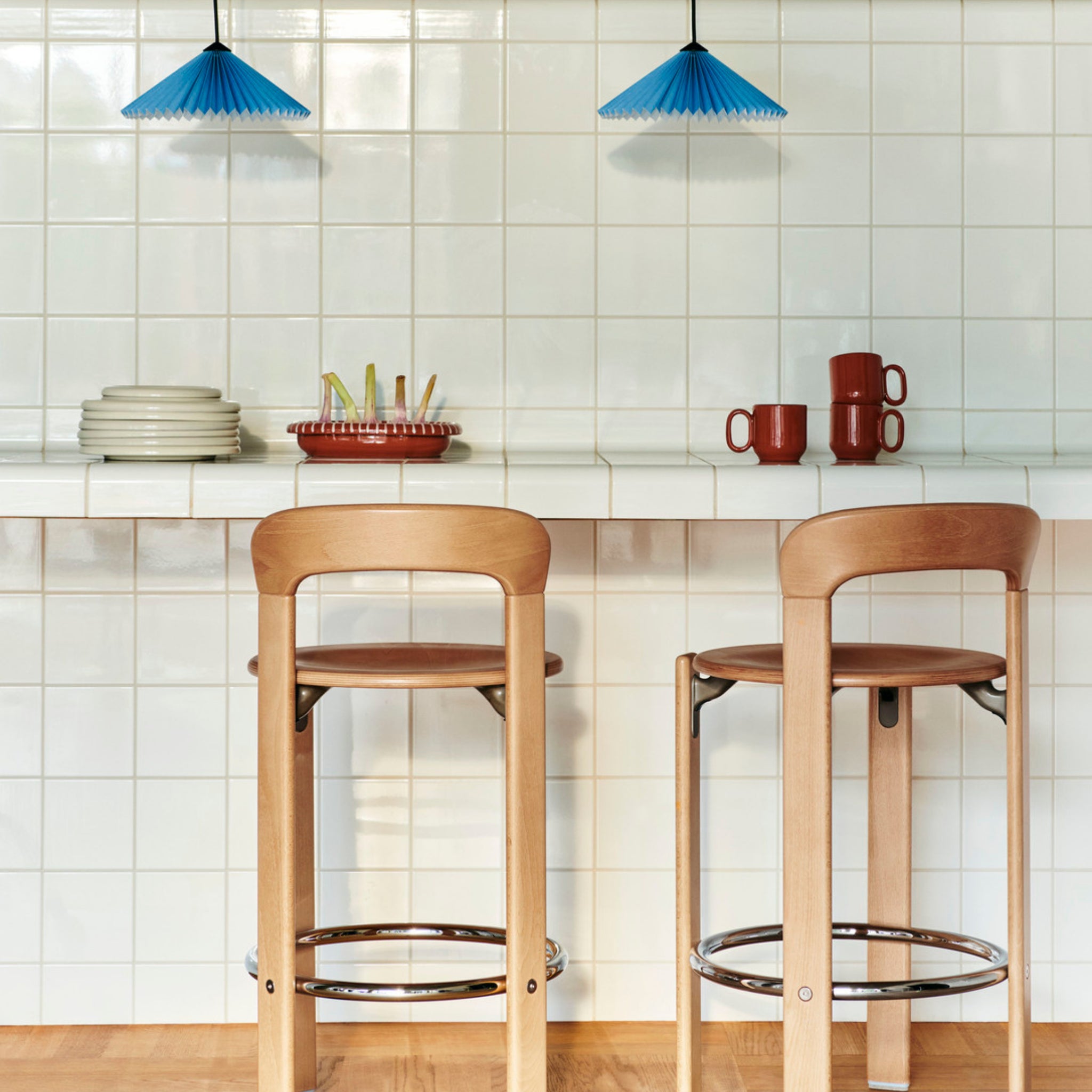 Een moderne keuken met twee houten barkrukken op een wit betegeld aanrecht, gestapelde borden, kopjes en keukengerei, waaronder de Hay Barro Ovale Schaal Terracotta 20/27 van Hay, met twee blauwe hanglampen boven je hoofd.