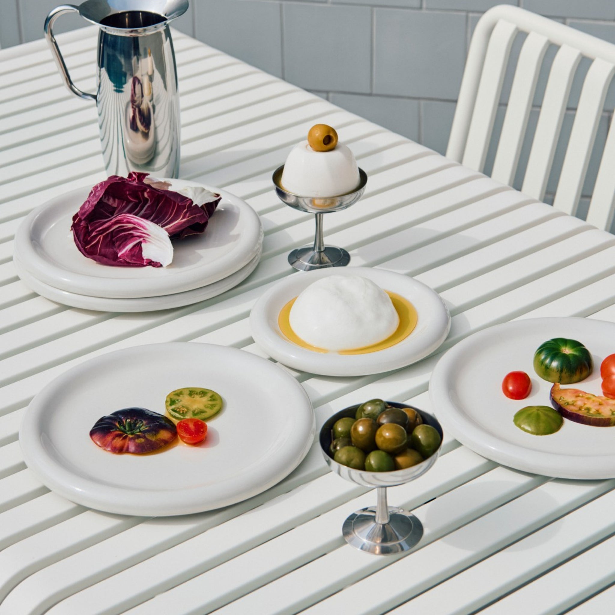 Een Hay Barro Borden Off-White D24 bordenset van Hay bevat borden met kleurrijke tomaten, paarse sla, groene olijven, een dessert met een olijf erop, een metalen kan en een schaal met wit schuim en gele saus tegen betegelde muren.