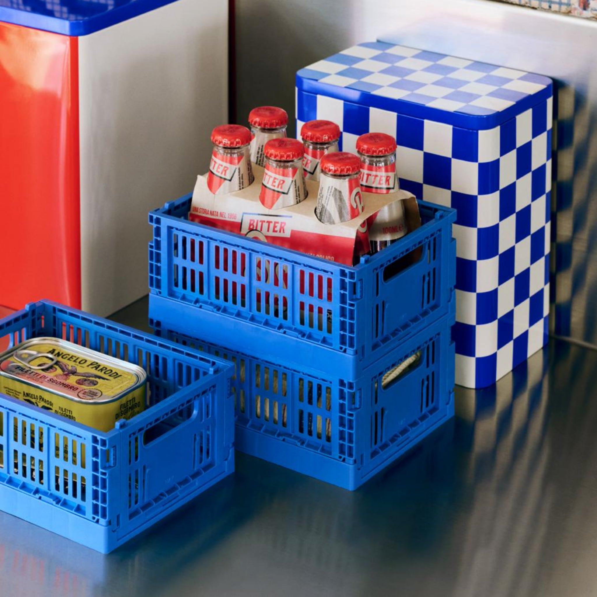 Twee blauwe plastic opbergdozen op een roestvrij stalen oppervlak bevatten drank in flessen met het label BUTTER en een geel blikje. Daarachter staan rode en witte bakjes, waaronder de Hay Blikken Doos Blauw Off-White L van Hay met droge etenswaren.