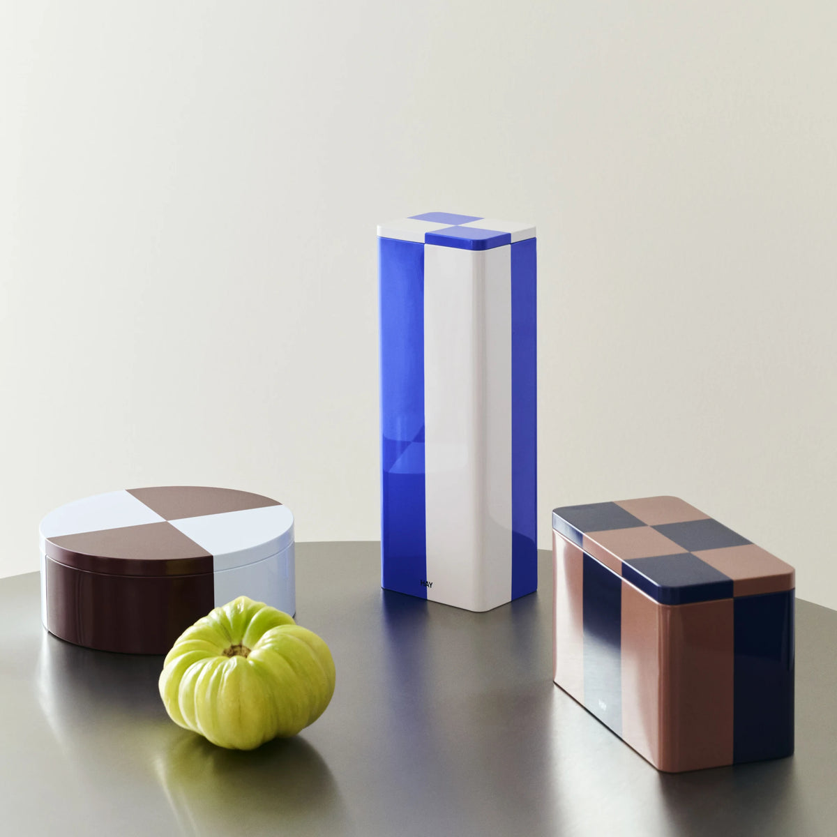 De Hay Blikken Doos Bordeaux Ijsblauw Rond op een donkere tafel naast een groene tomaat, met een blauw-witte en een geblokte blikken doos.