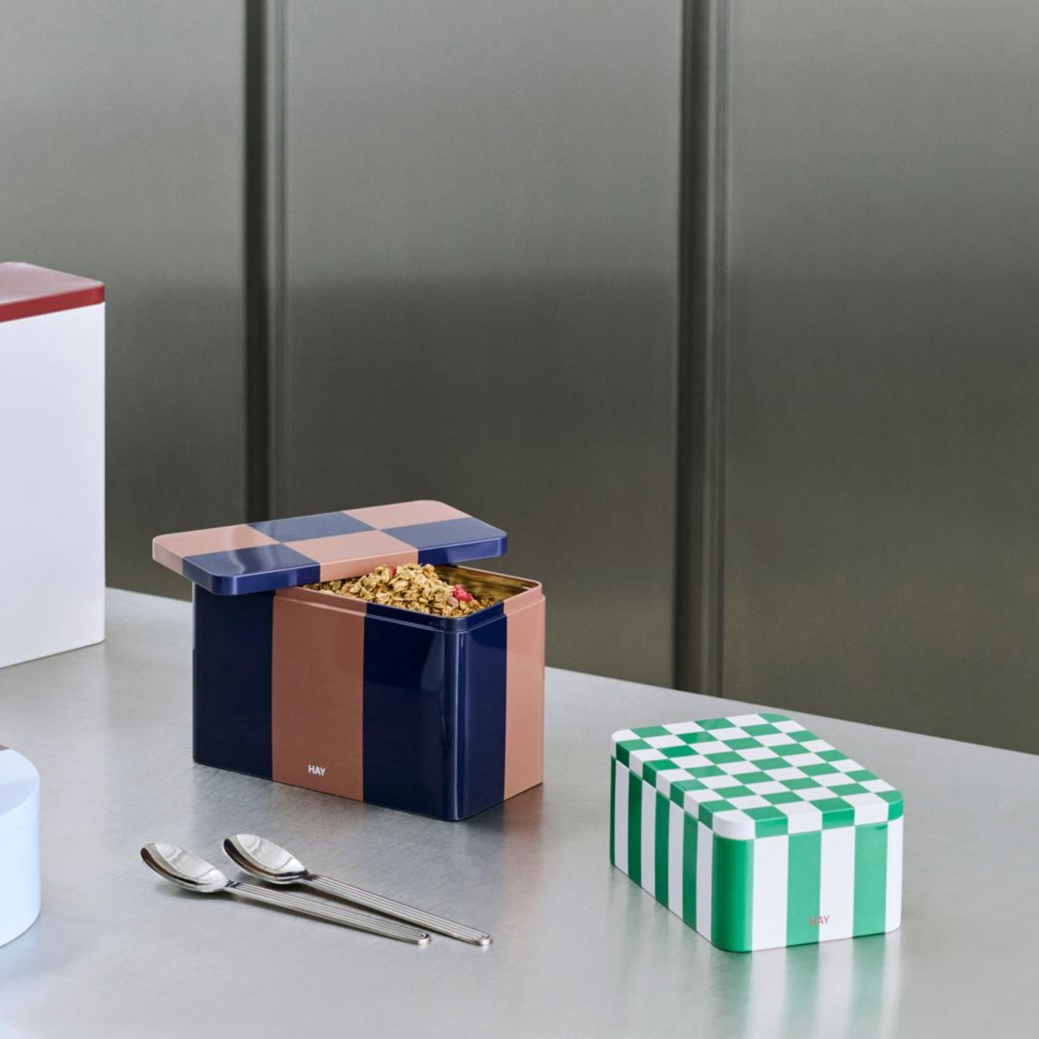 Een open rechthoekig blik met een roze en marineblauw geruit deksel onthult ontbijtgranen, terwijl de Hay Blikken Doos Groen Off-White S van Hay, met een groen dambordmotief, vlakbij staat. Twee lepels rusten op het metalen aanrecht.