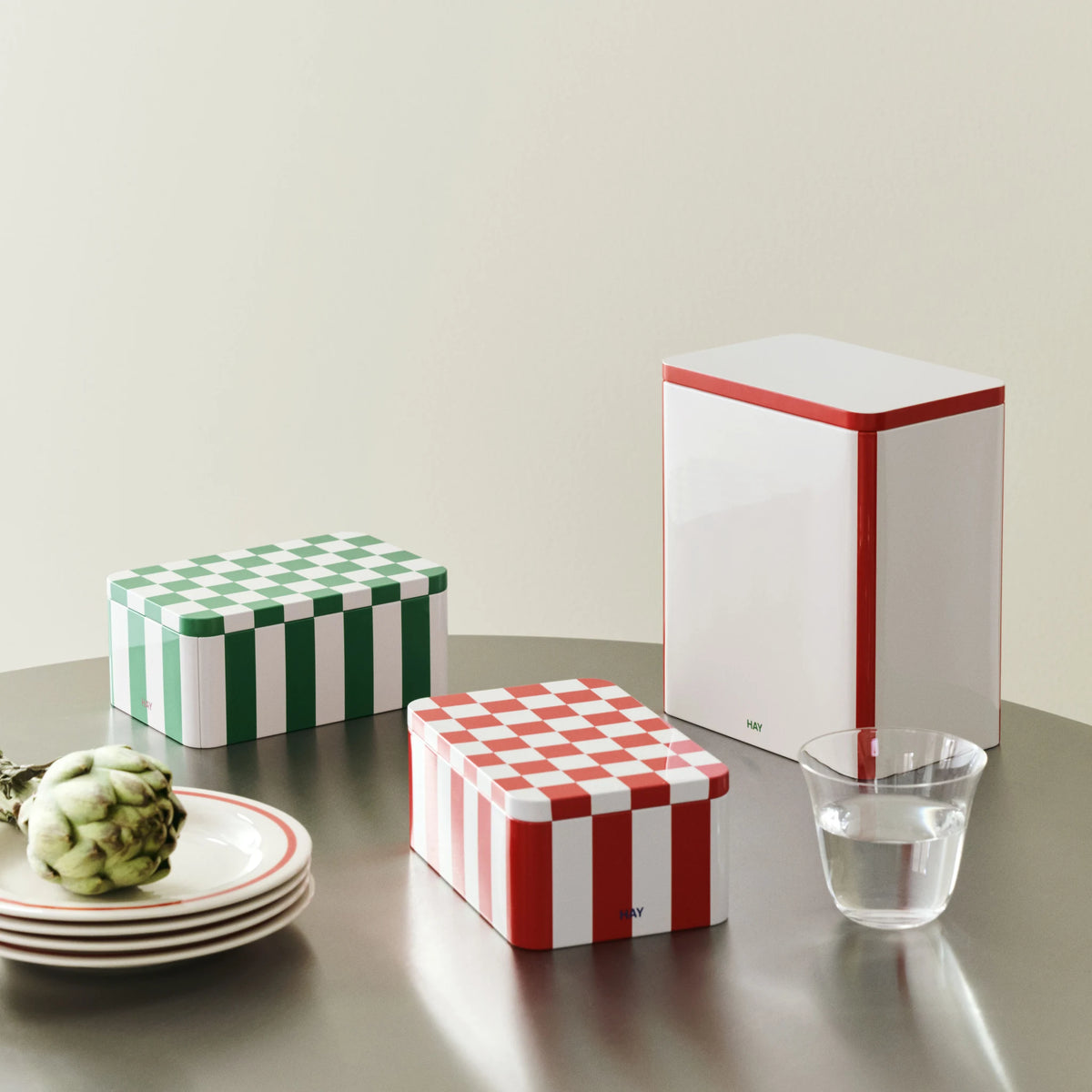De Hay Blikken Doos Rood Off-White S met rood-witte strepen op een ronde tafel, naast een groen-witte blikken doos, een hoge blikken doos en een artisjok.