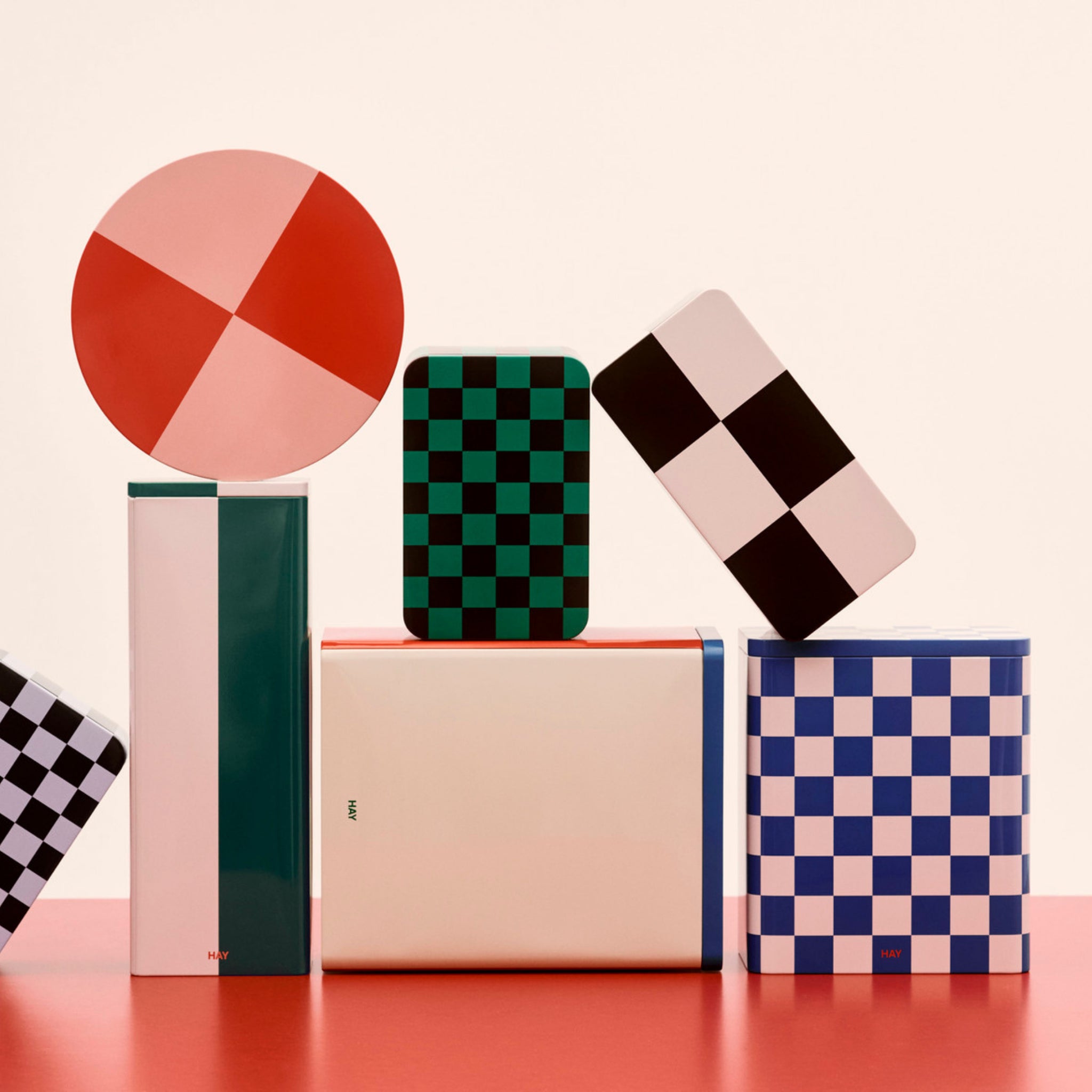 De Hay Blikken Doos Blauw Off-White L van Hay is een kleurrijk geometrisch opbergblik met blauwe en gebroken witte patronen, speels gestapeld op een rood oppervlak tegen een lichte achtergrond.