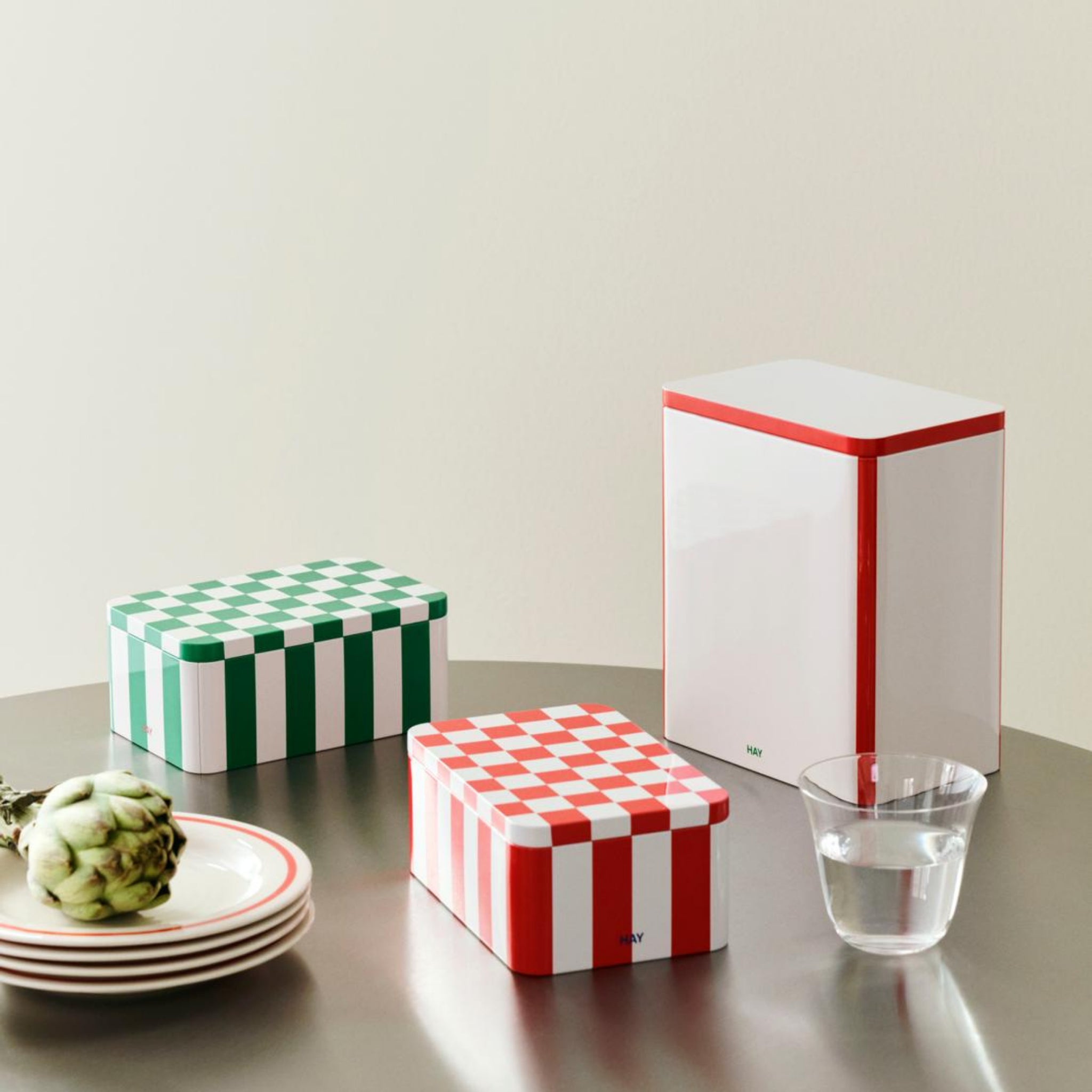 Drie Hay Blikken Doos Off-White Rood L containers met dambordmotieven, twee rood-wit en een groen-wit, staan op een tafel met borden, een artisjok en een glas water tegen een neutrale achtergrond.