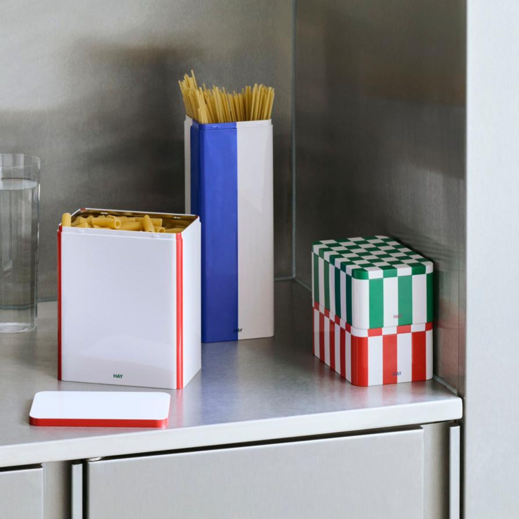 Op een roestvrijstalen aanrecht staat de Hay Blikken Doos Off-White Rood L container van Hay met kleurrijke patronen, sommige gevuld met droge pasta, naast een glas water in een moderne keuken.