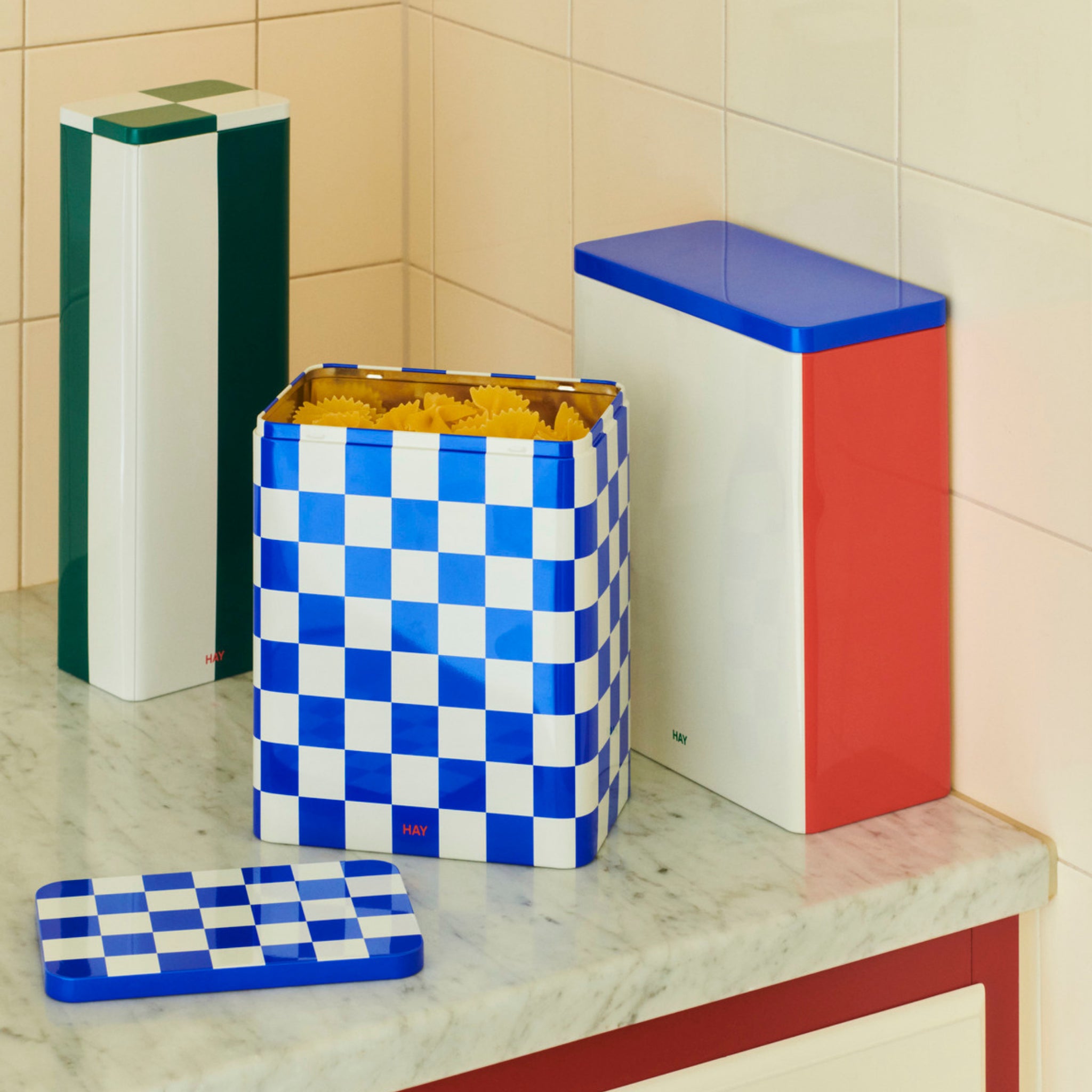 Drie kleurrijke Hay opbergdozen staan op een marmeren aanrecht. De voorste, de Hay Blikken Doos Blauw Off-White L, heeft een blauw en gebroken wit geruit ontwerp en bevat pasta-perfect voor het bewaren van droge etenswaren.