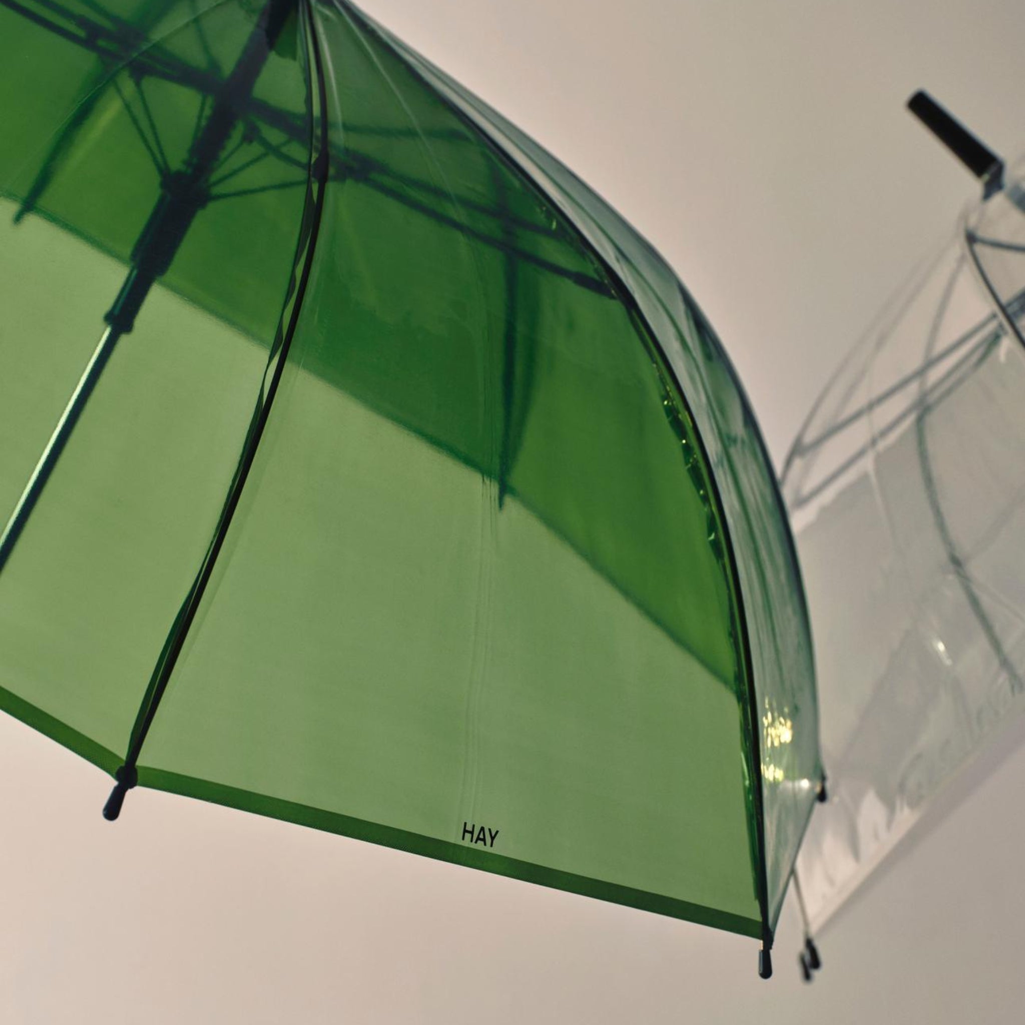 Een close-up van de Hay Canopy Paraplu Groen, met het HAY-logo, zit naast een transparante paraplu, beide vastgelegd tegen een eenvoudige, lichte achtergrond.