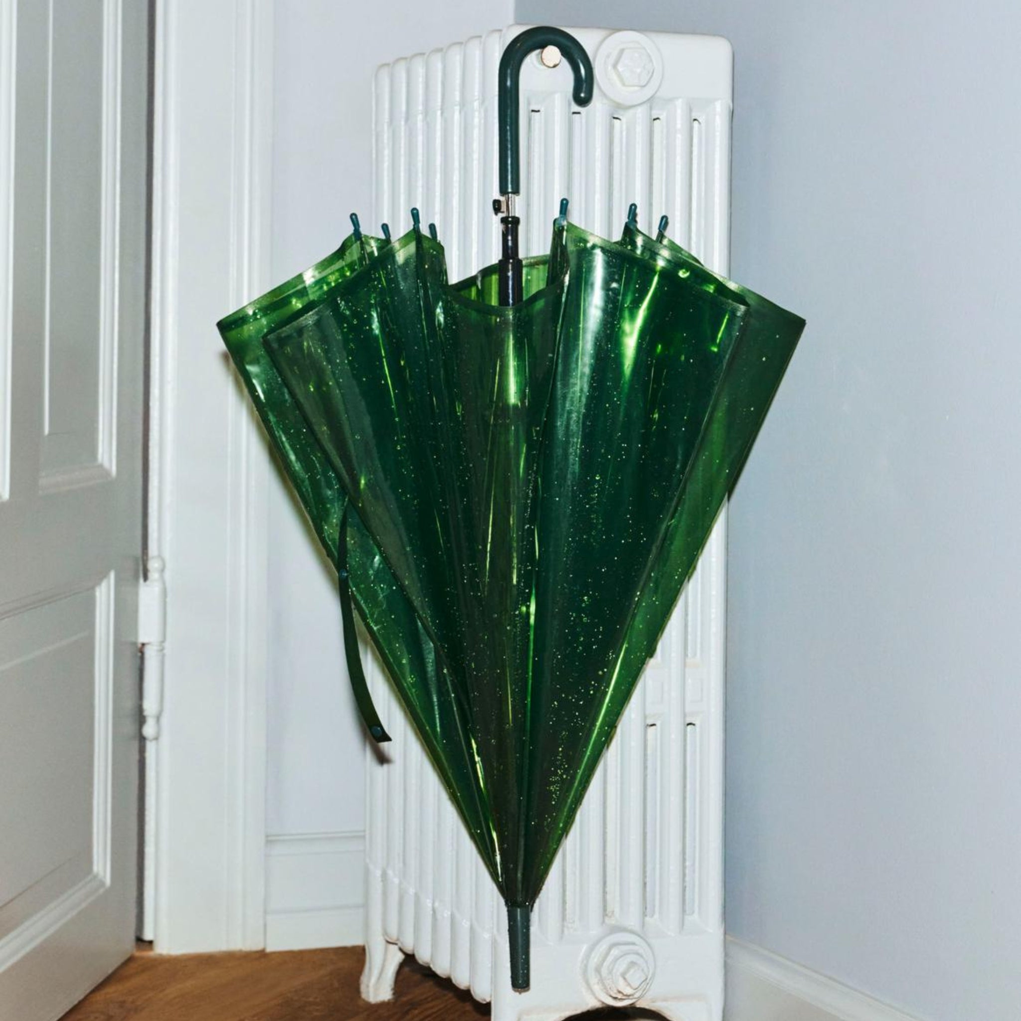 De Hay Canopy Paraplu Groen van het merk Hay, gemaakt van gerecycled materiaal en voorzien van een gebogen handvat, staat gesloten tegen een witte radiator in de hoek van een lichte kamer met houten vloer.