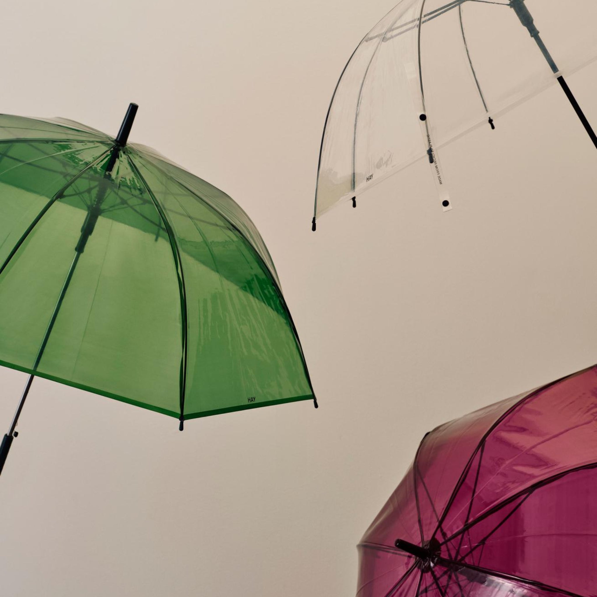 De Hay Canopy Paraplu Groen van Hay, gemaakt van gerecycled materiaal, wordt links getoond tegen een beige achtergrond met roze en heldere paraplu's rechtsonder en boven.
