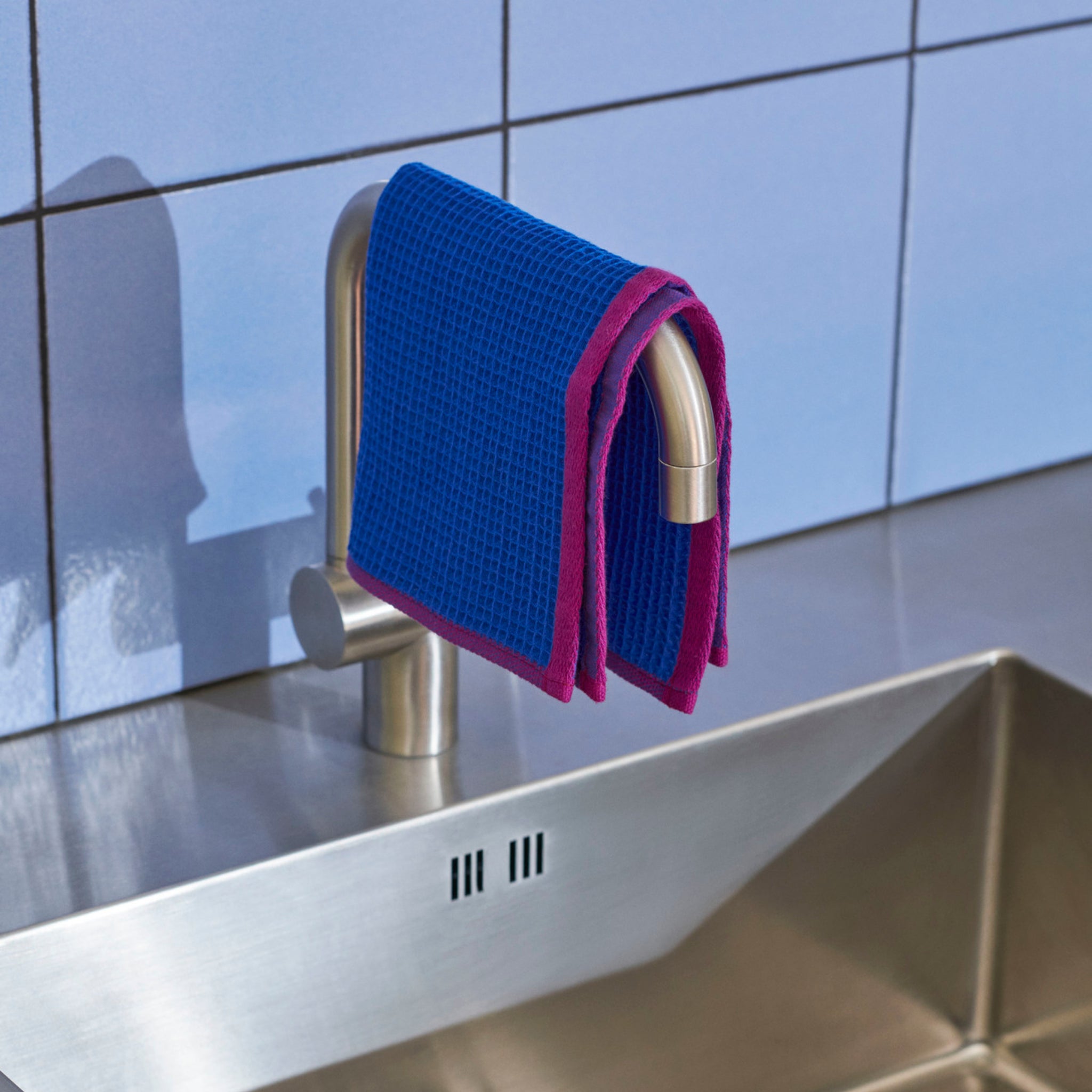 Een Hay Canteen Vaatdoek Blauw Geel van Hay is gedrapeerd over een zilveren kraan boven een roestvrijstalen gootsteen, tegen een lichtblauw betegelde muur - ideaal voor efficiënt keuken schoonmaken.
