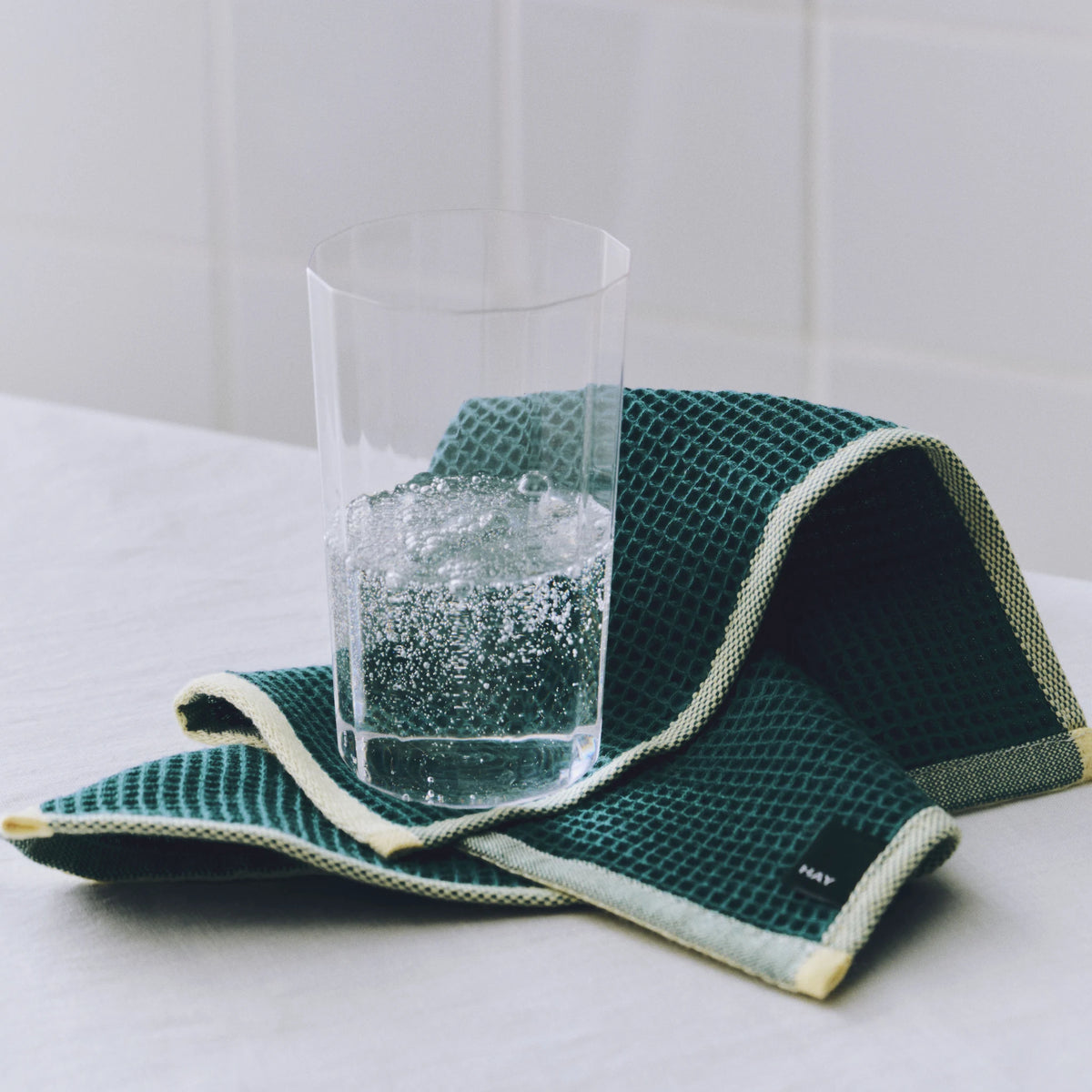 Een groene Hay Canteen Vaatdoek uit de set Geel Groen met wafelstructuur en gele rand, onder een glas water op een witte tafel.