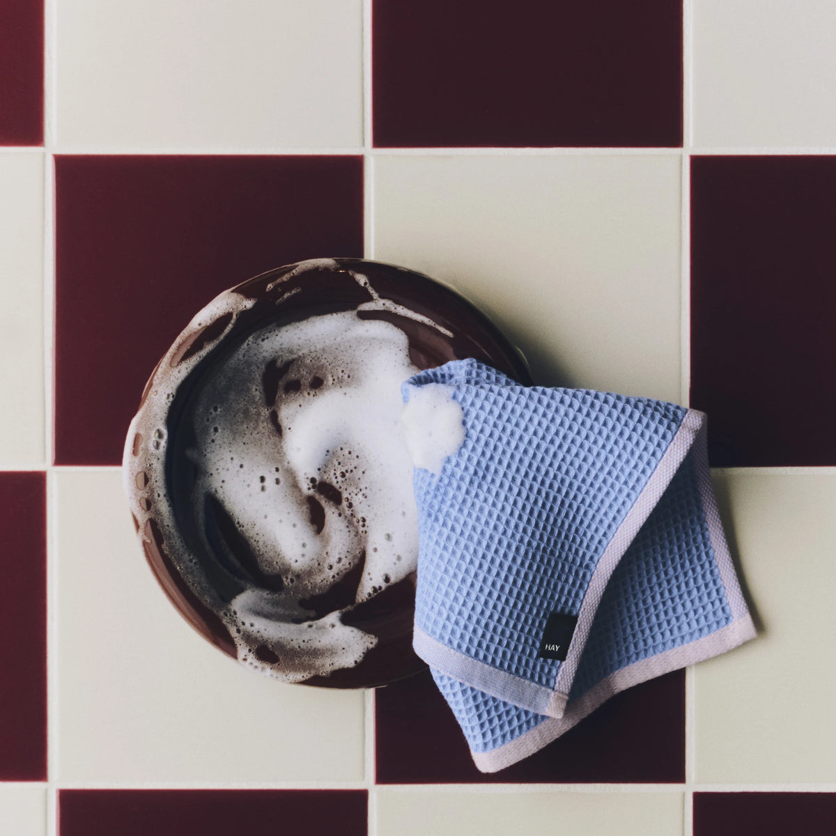Een hemelsblauwe Hay Canteen Vaatdoek met roze rand naast een kom met schuim, op een geblokte bordeaux-crème vloer.
