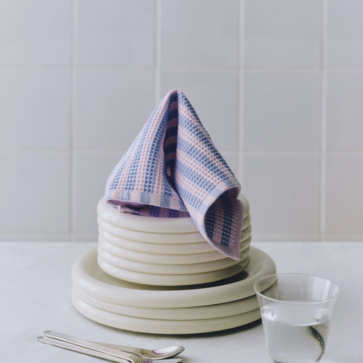 De gestreepte Hay Canteen Vaatdoek uit de set Hemelsblauw Roze ligt over een stapel crèmekleurige borden, naast een glas en lepels.
