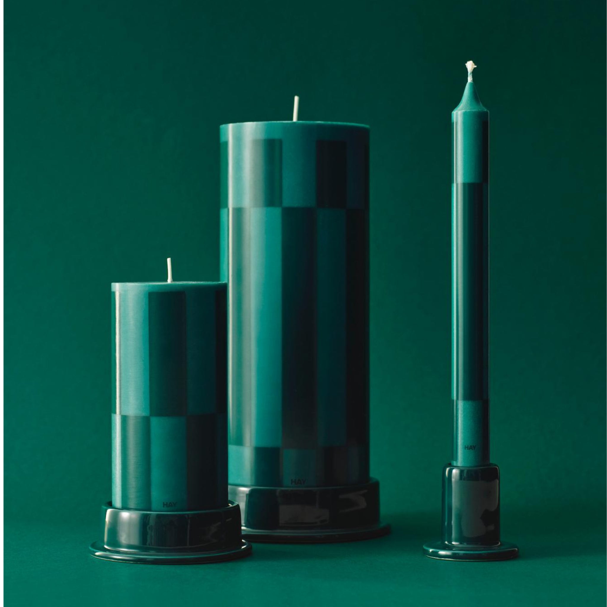 Drie groene kaarsen van verschillende hoogtes en vormen, waaronder de Hay Column Kaars Donkergroen H25 van Hay, rusten op bijpassende houders tegen een effen groene achtergrond. Elke sierkaars heeft een subtiel dambordpatroon voor extra stijl.