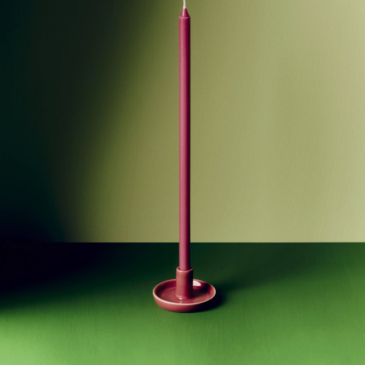 De Hay Disc Kandelaar Bessenroze XS met een lange bordeauxrode kaars, op een donkergroen oppervlak tegen een olijfgroene muur.