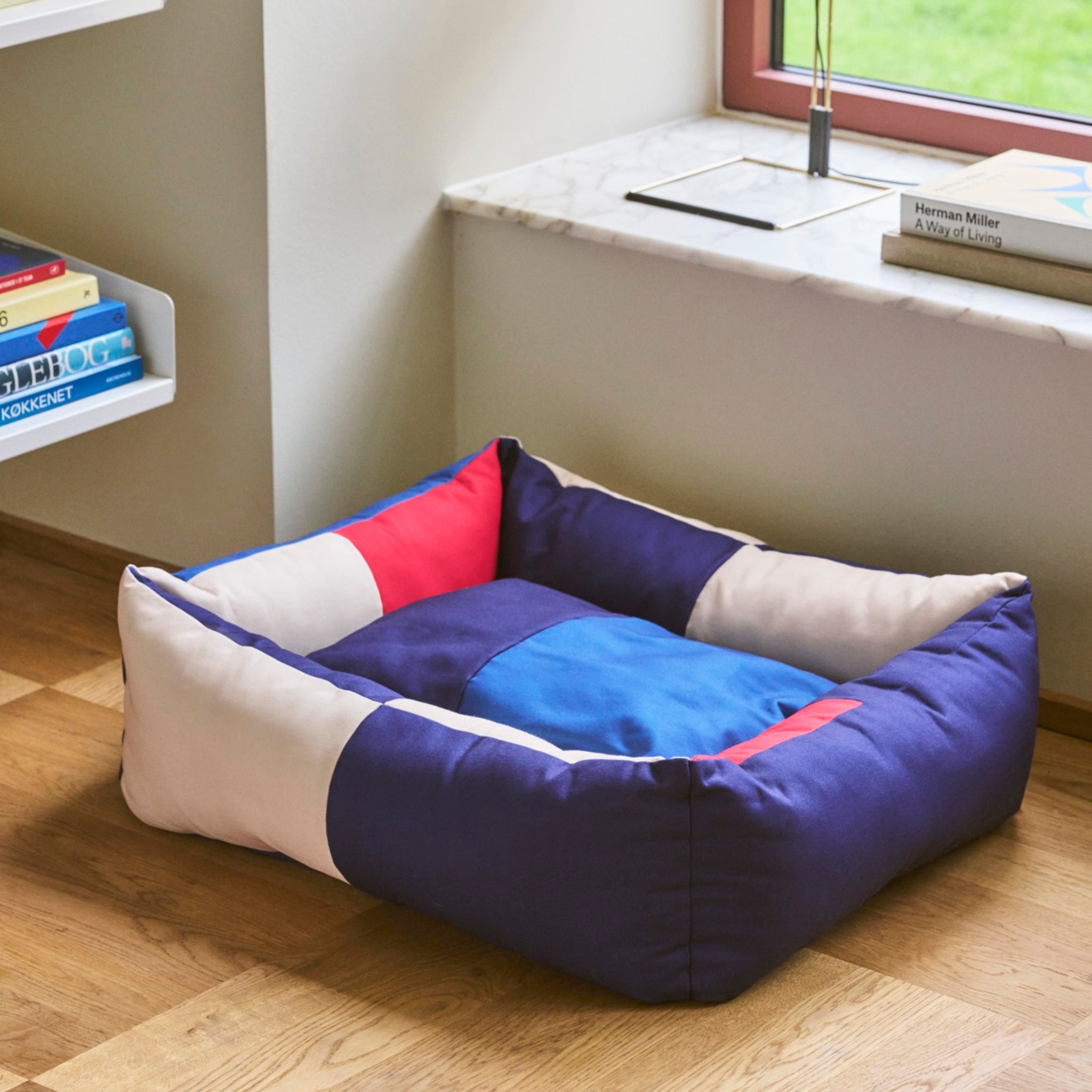 De Hay Hondenmand Rood Blauw 65/78 van Hay, met zijn rode en blauwe vlekken, staat op een houten vloer bij een raam en een boekenplank.