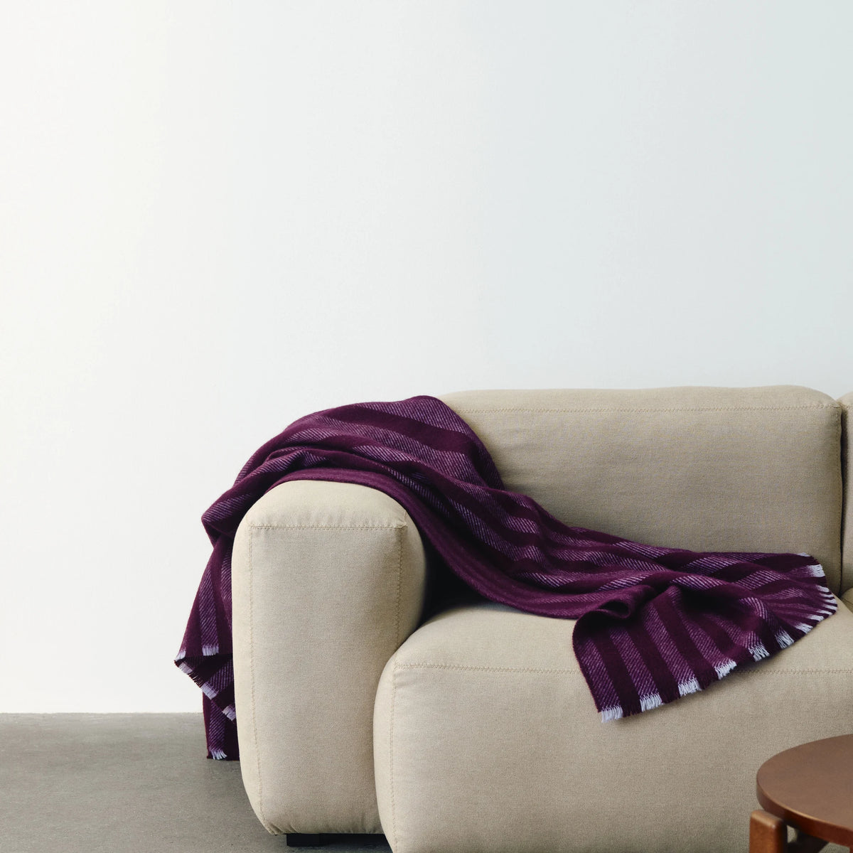 De Hay Duo Plaid Pruim ligt losjes gedrapeerd over de armleuning van een beige sofa in een lichte, rustige woonkamer, met een houten bijzettafel op de voorgrond.
