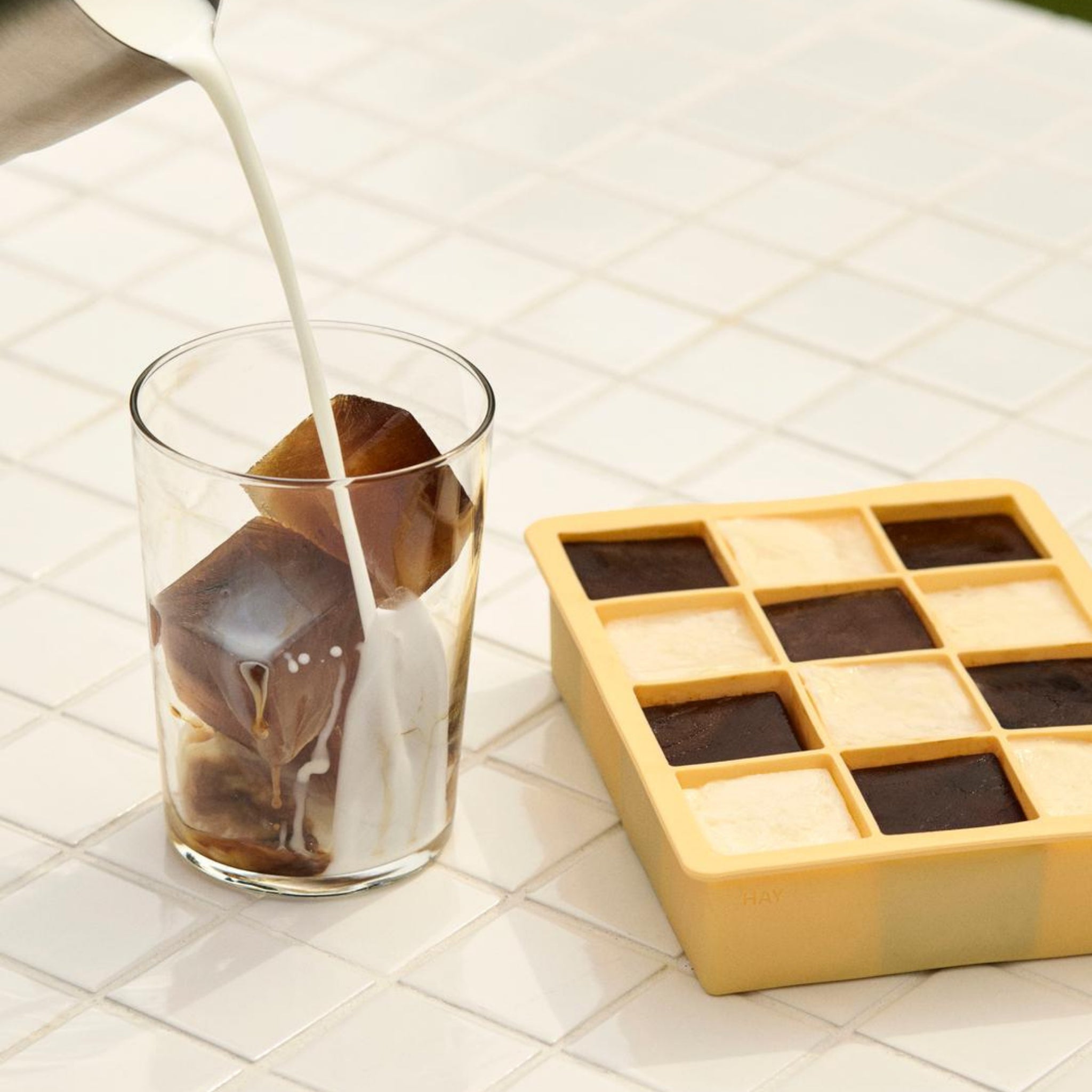 In een glas met koffie-ijsblokjes is melk gegoten. Ernaast staat een Hay ijsblokjesvorm Vierkant XL Blauw van Hay, gevuld met koffie- en melkijsblokjes, alles op een wit betegeld oppervlak.