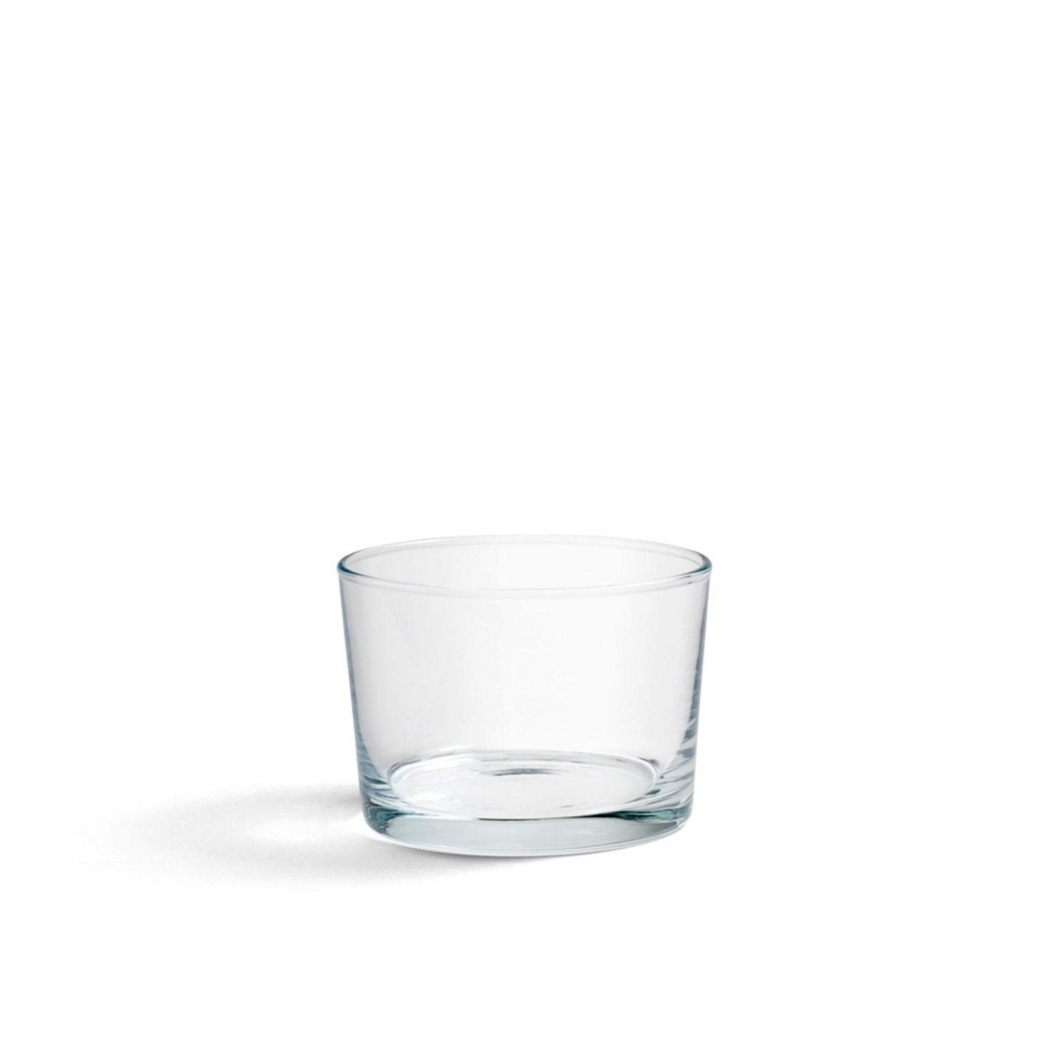 De Hay Glas Helder S van Hay is een minimalistische glazen beker met rechte zijkanten, gecentreerd op een effen witte achtergrond en met een vage schaduw. Dit kleine glas heeft een stapelbaar, ruimtebesparend ontwerp.
