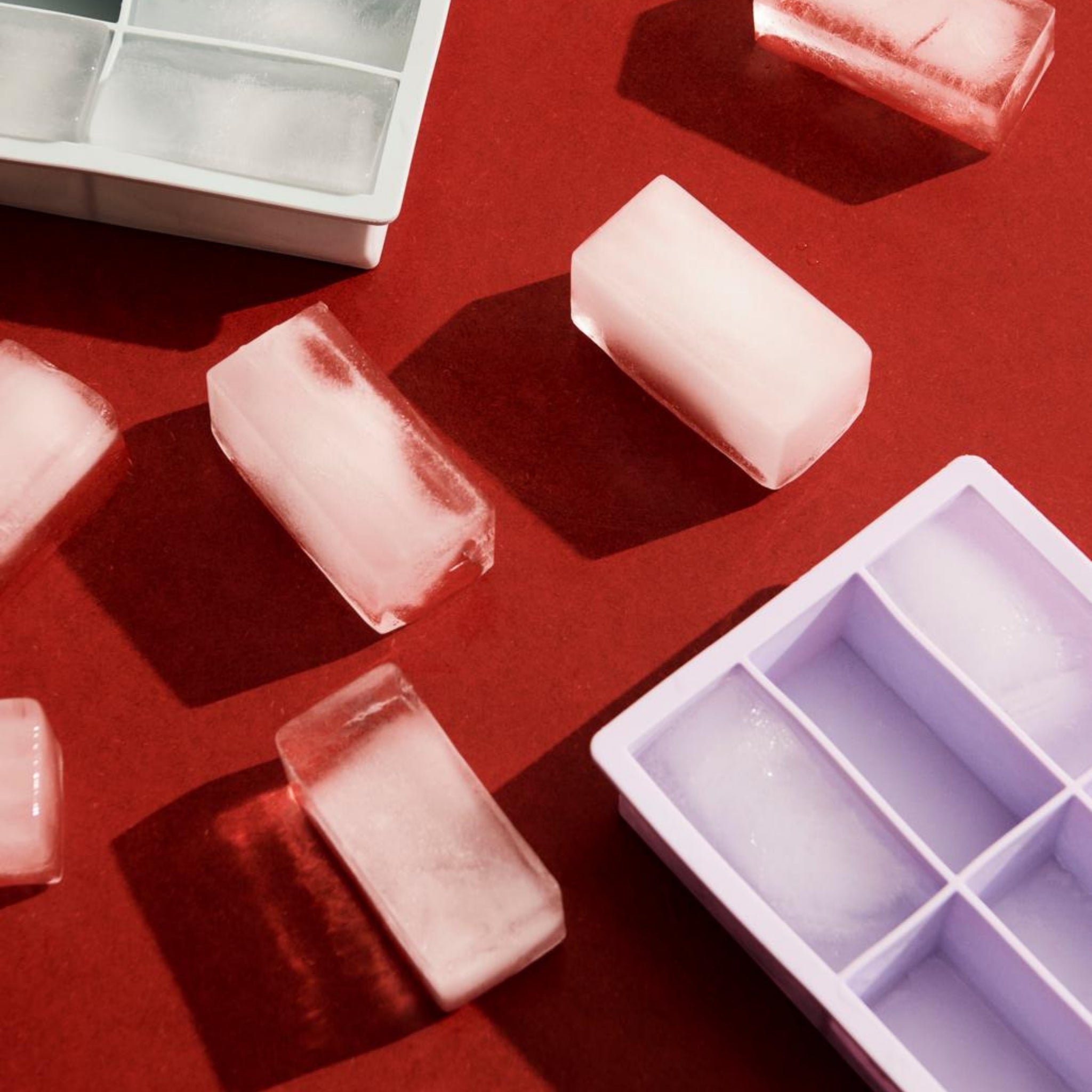 De Hay ijsblokjesvorm Rechthoekig XXL Ijsblauw ijsblokjes worden weergegeven op een rood oppervlak naast witte en paarse siliconen bakjes, met schaduwen die duiden op heldere, directe verlichting.
