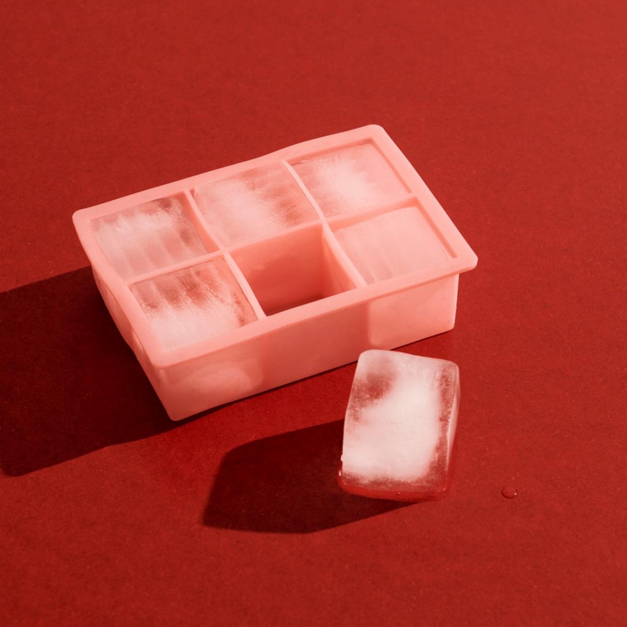 Een Hay ijsblokjesvorm Vierkant XXL Roze van Hay, gevuld met meerdere XXL ijsblokjes, staat op een rood vlak met één blokje ernaast.