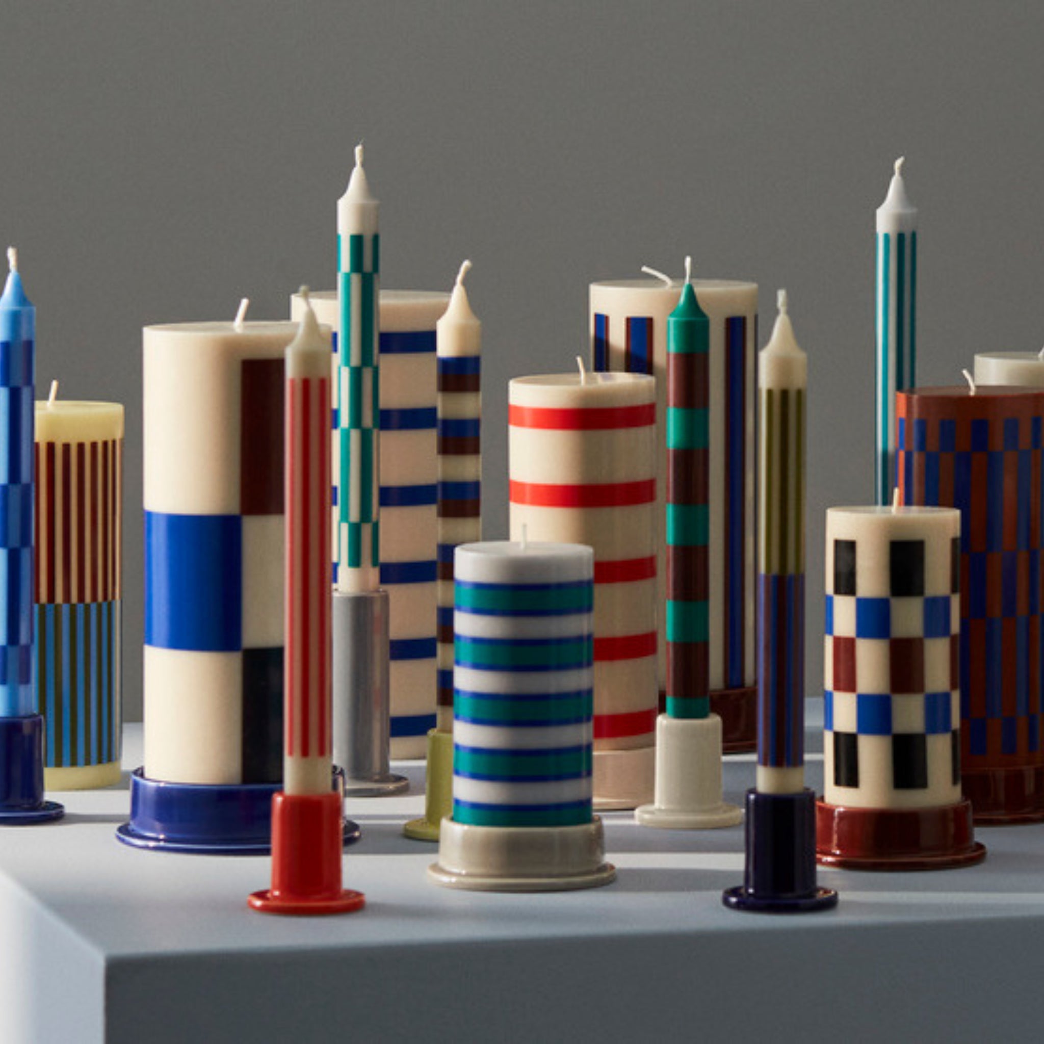 Kleurrijke kaarsen met geometrische patronen en blauwe, rode, groene, beige en witte strepen en ruiten staan op een grijs oppervlak naast de Hay Tube Kandelaar Bordeaux kandelaar van Hay.