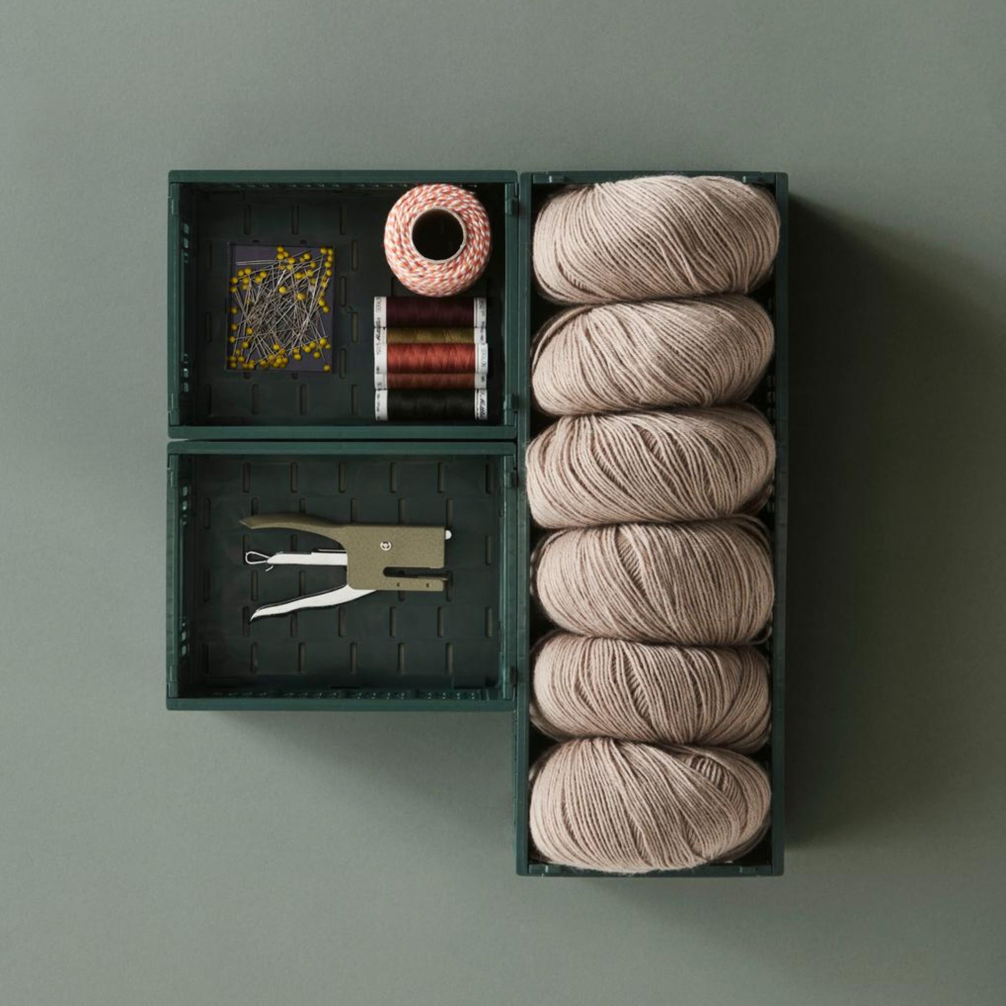 In drie Hay Kleur Krat Mini Donkergroen dozen van Hay, gemaakt van gerecycled plastic, kunnen garen, garenklosjes, gele spelden, touw en een nietmachine netjes worden opgeborgen op een bijpassende donkergroene achtergrond.