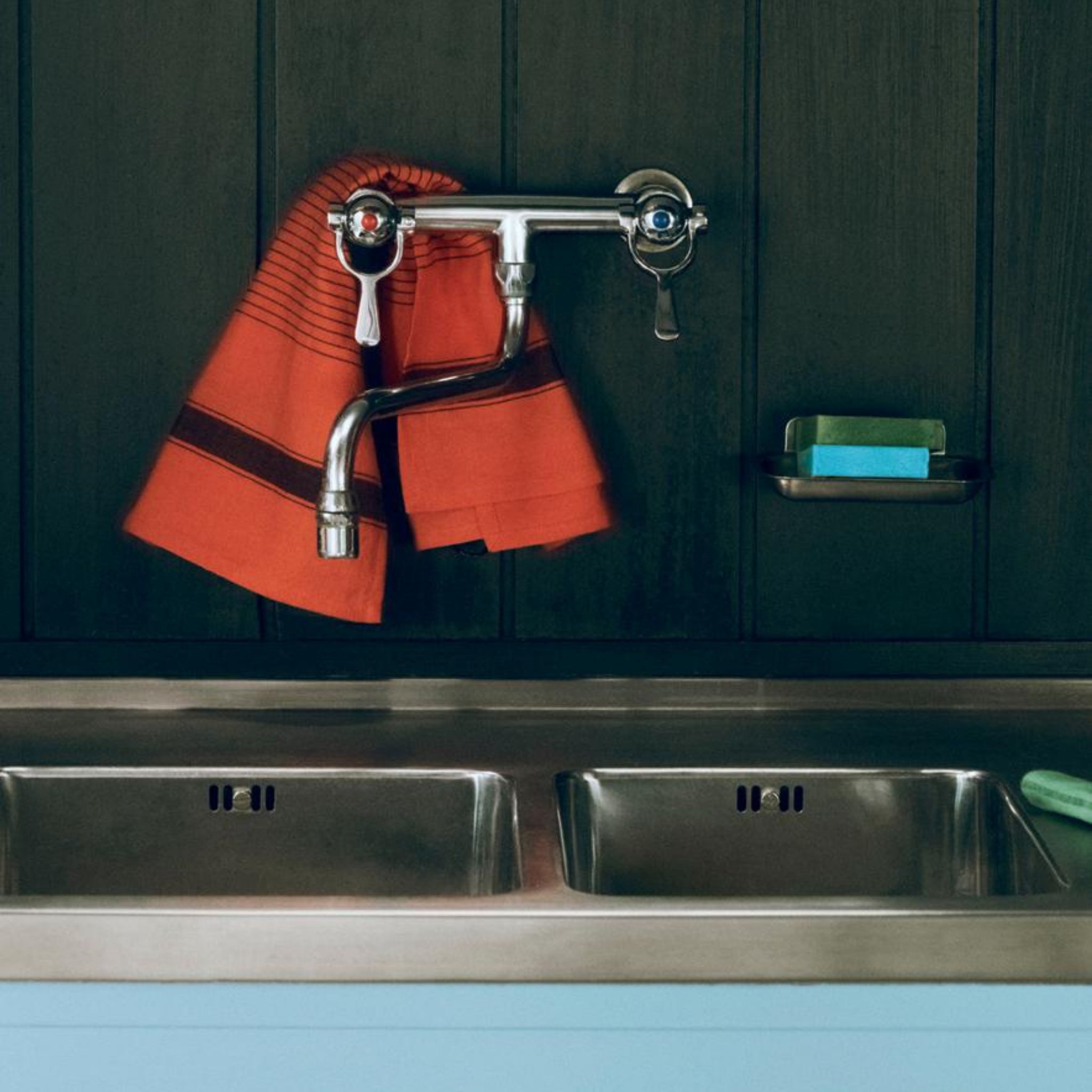 Een aanrecht met twee waskommen, een zilveren kraan en een Hay Linear Theedoek Rood die boven de kraan hangt. Een blauwe spons en groen schuursponsje rusten op een plankje boven de gootsteen tegen een donkere muur.