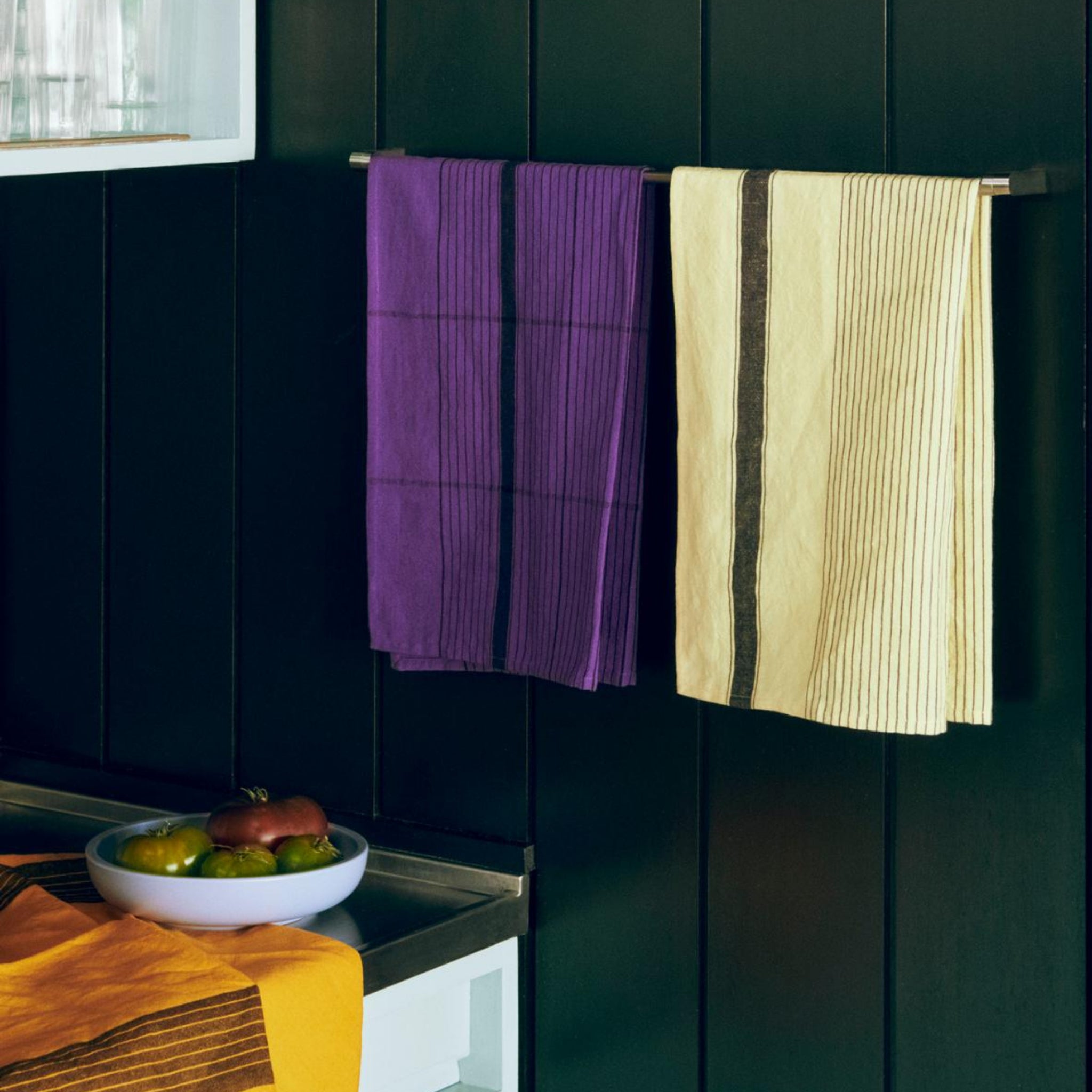 Twee handdoeken - een paarse Hay Linear Theedoek en een crèmekleurig gestreepte linnen-katoenen handdoek - hangen naast elkaar aan een metalen stang. Eronder liggen verschillende groenten in een schaal op een aanrecht dat bedekt is met de gele Hay Linear Theedoek.