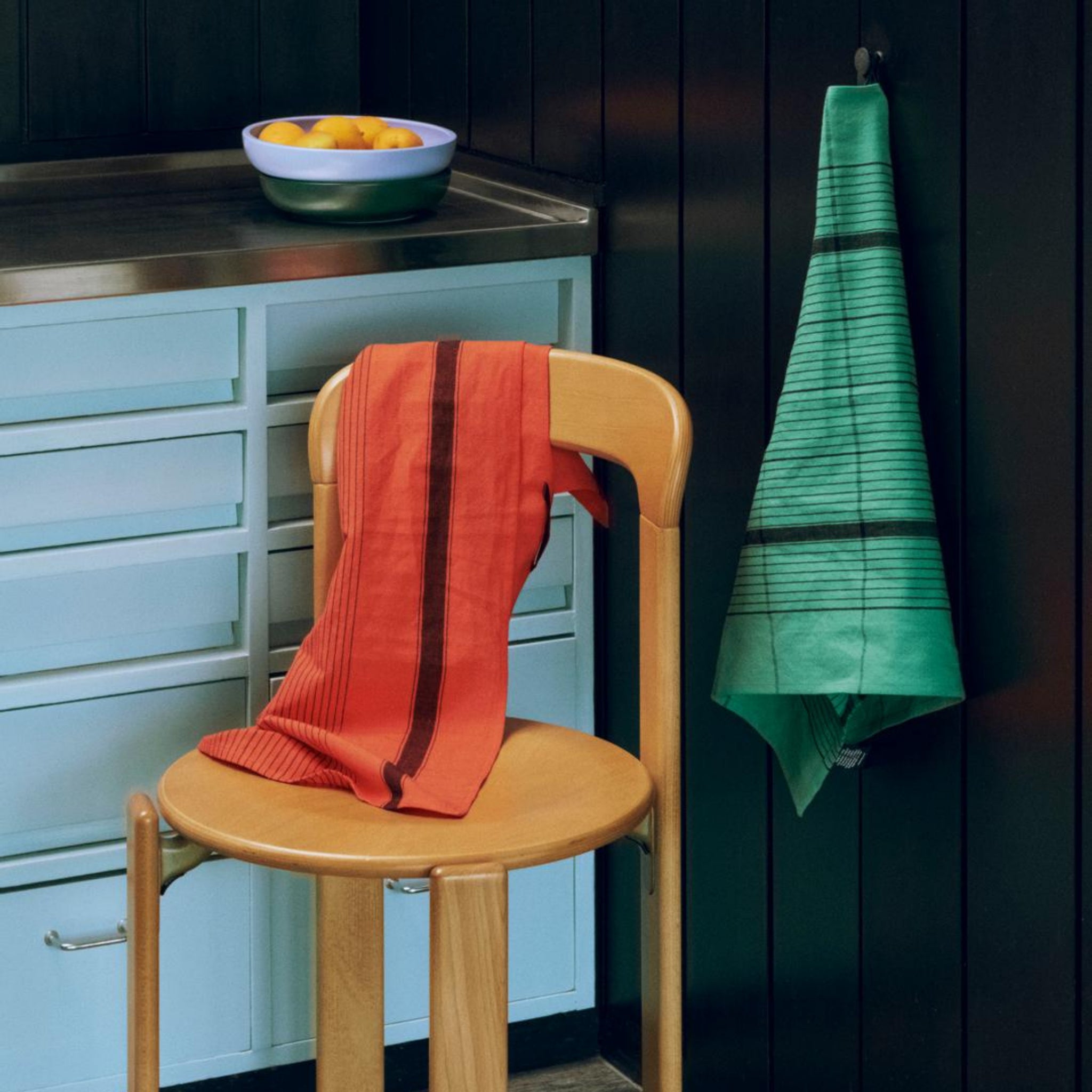 Een Theedoek Rood van Hay Linear is over een houten stoel gedrapeerd, terwijl een groene handdoek aan een haakje in de keuken hangt, met sinaasappels in een schaal op het roestvrijstalen aanrecht erachter.