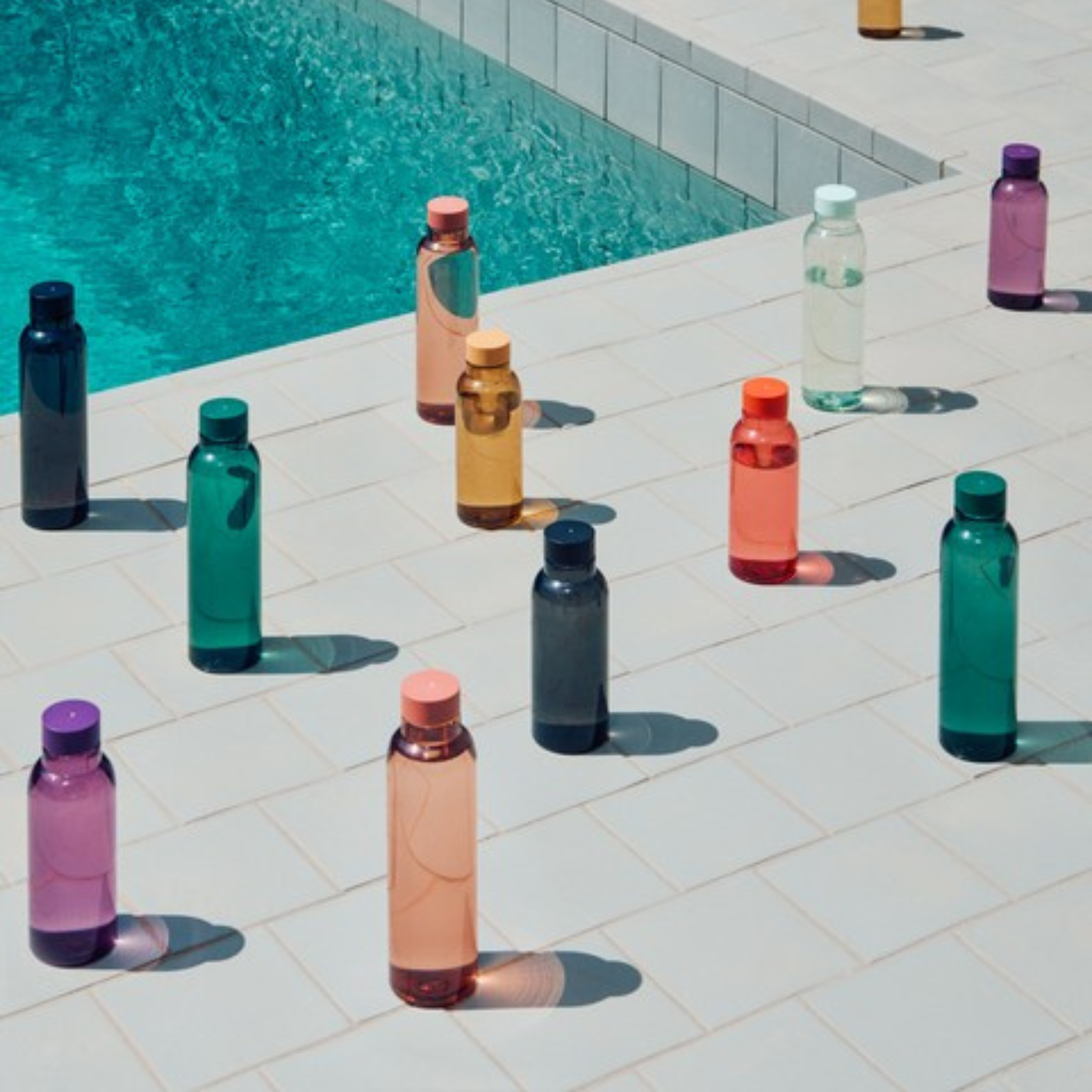 Hay Miz Waterfles Houtskool 720 ml van Hay staan rechtop op lichte tegels aan de rand van een zwembad, waarbij elke fles een schaduw werpt in het zonlicht.