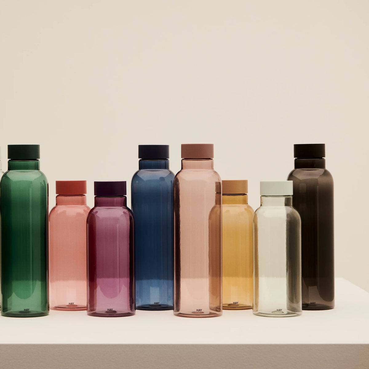 Een rij Hay Miz waterflessen in verschillende kleuren en maten op een witte sokkel, waaronder de Hay Miz Waterfles Karamel 540 ml.