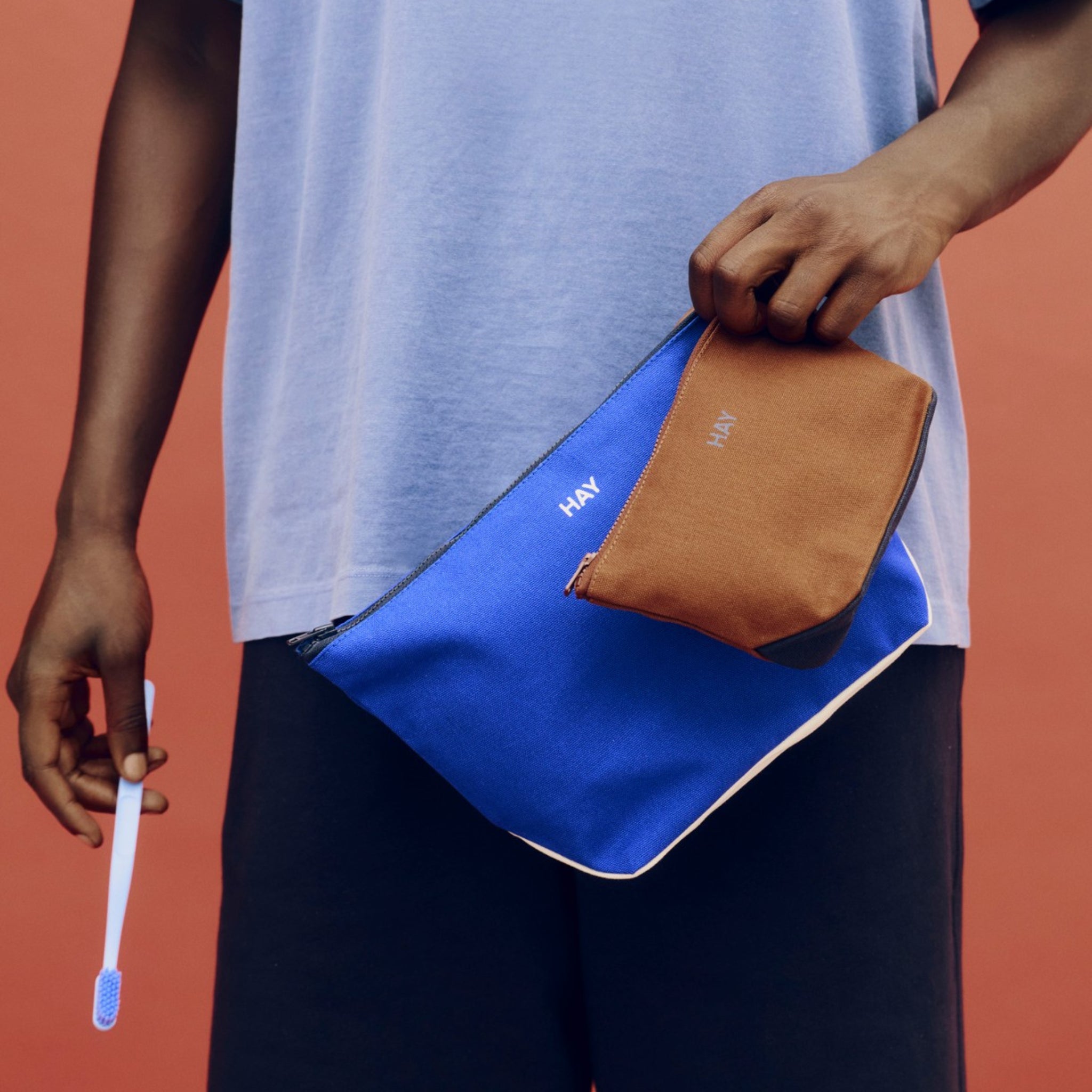 Een persoon in een lichtblauw shirt houdt een Hay Multi Opbergzakje Elektrisch Blauw M en een bruin tasje in de ene hand, en een blauwe tandenborstel in de andere, staande voor een rood-oranje achtergrond die Scandinavisch design accentueert.