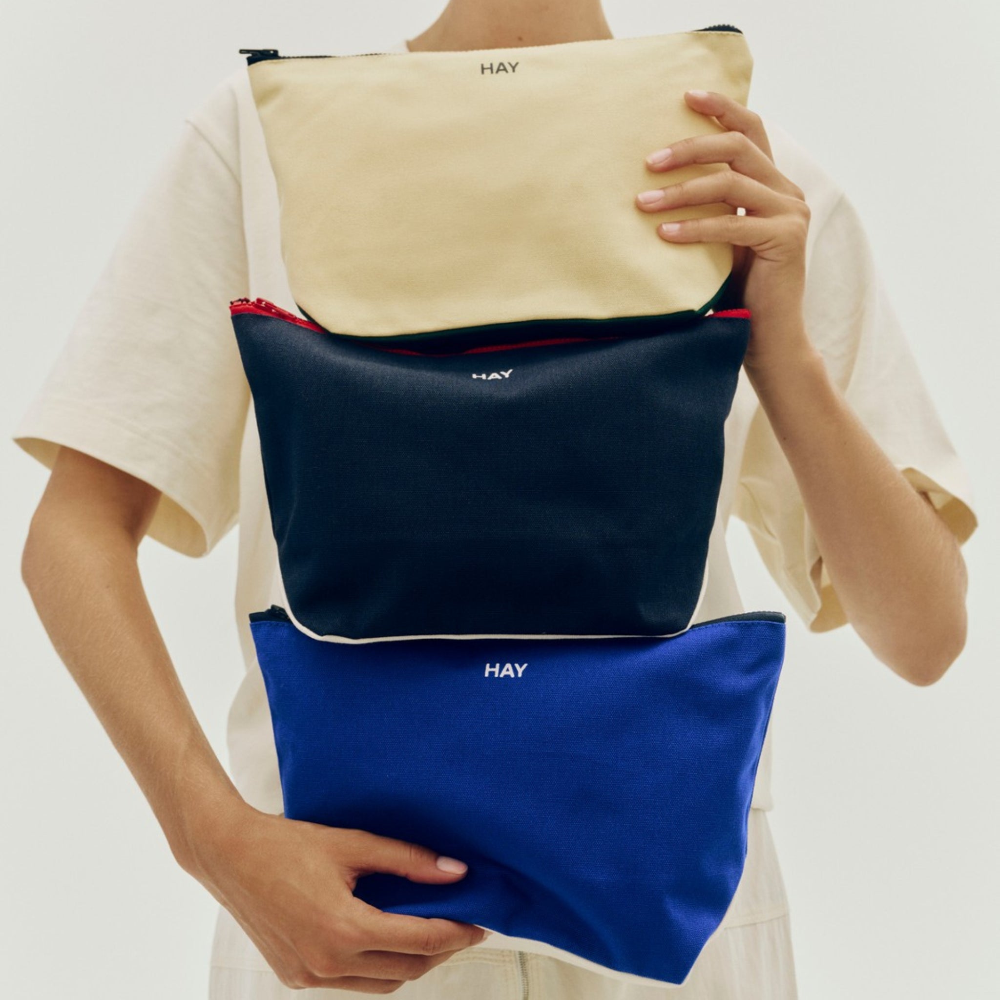 Een persoon in een beige overhemd houdt drie Hay Multi Opbergzakjes vast-cream, navy en Midnight Blauw M van Hay, verticaal gestapeld om een stijlvolle opbergoplossing te laten zien tegen een effen lichte achtergrond.
