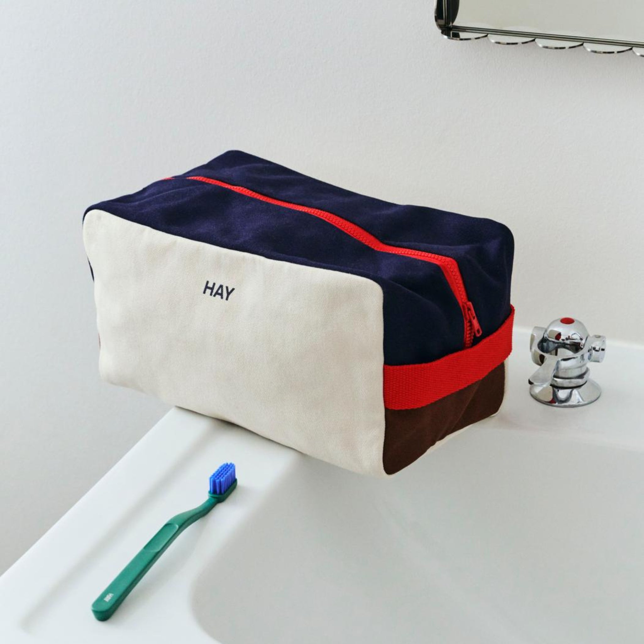 Een Hay Multi Toilettas Medium Crème van Hay, gemaakt van biologisch katoen met navy, crème, bruine en rode kleuren en een rode rits, staat op een witte wastafel. Voorop staat een blauwe tandenborstel met een groen handvat.
